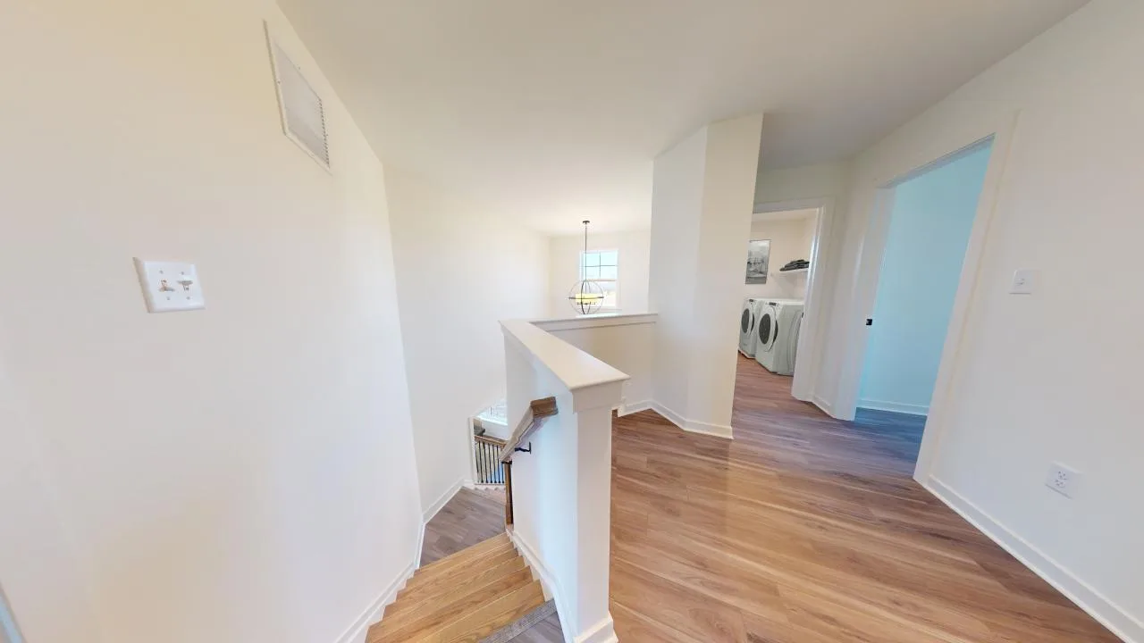 A bright hallway with light wooden floors, leading to a laundry room with washer and dryer. A stairway with white railing goes down, and a room with blue walls is visible on the right. Natural light fills the space.