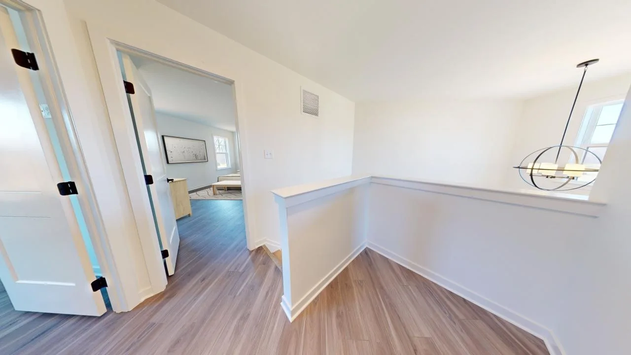 A bright hallway with light wood floors, white walls, and a half wall railing. An open doorway leads to a room with blue flooring and framed artwork, and a modern light fixture hangs in the background.