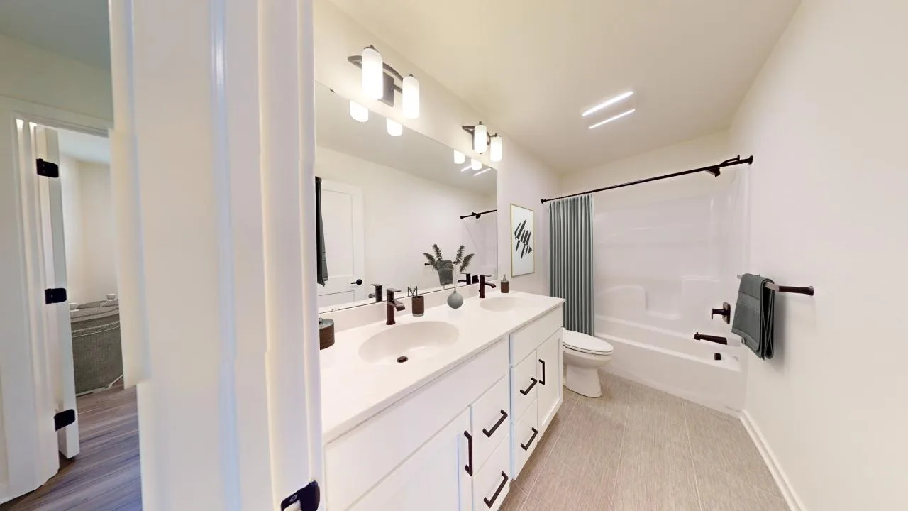 Modern bathroom with white walls, double-sink vanity, large mirror, bathtub with shower and dark fixtures, towel on rack, and a decorative plant near the sinks. Doorway leads to another room with wood flooring.