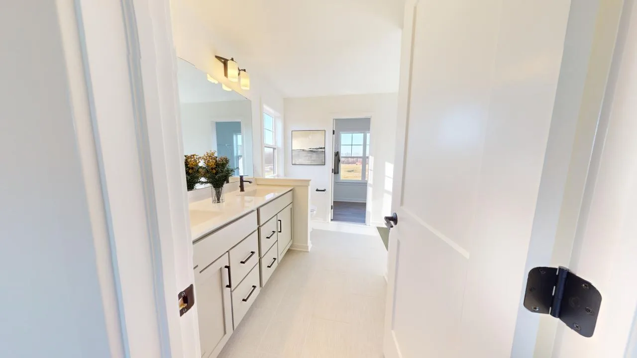 A bright, modern bathroom with white cabinets, a large mirror, a countertop vase with flowers, and an open door leading to a sunny room with artwork on the wall.