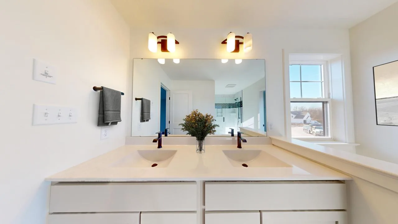 Modern bathroom with double sinks, a large mirror, two wall-mounted lights, gray towels on the left, a vase with greenery on the countertop, and a window letting in natural light on the right.