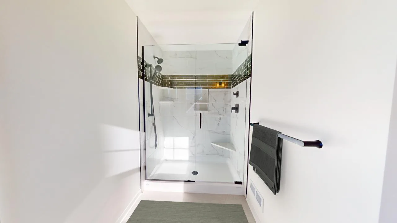 A modern walk-in shower with glass doors, marble-patterned walls, a built-in shelf, a black towel rack with a dark towel, and a light-colored floor mat in a bright, minimal bathroom.