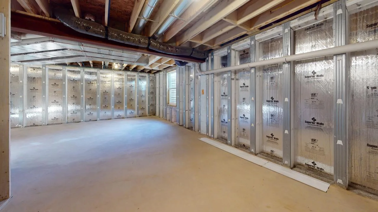 An unfinished basement with exposed ceiling beams, ductwork, and insulated walls covered in reflective material. The floor is bare concrete, and natural light enters through a small window.