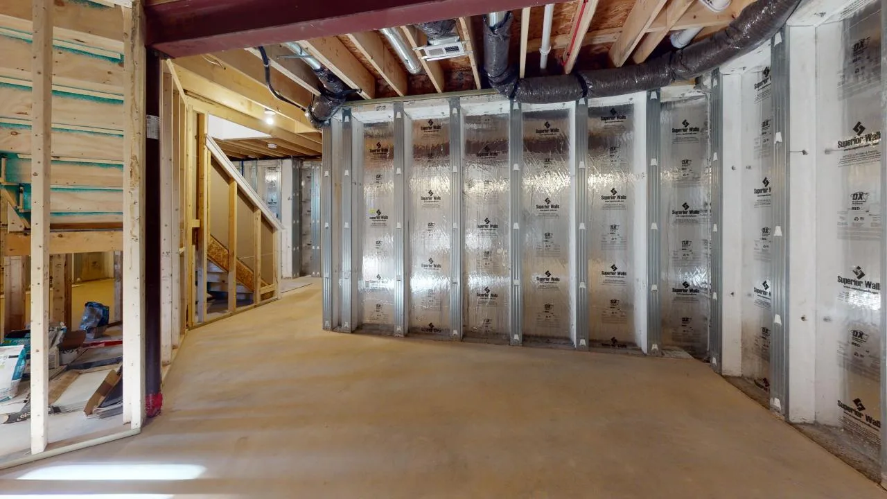 Unfinished basement with exposed wooden studs, foil insulation on some walls, exposed ceiling beams, ducts, and pipes. Concrete floor and a partially built staircase are visible.
