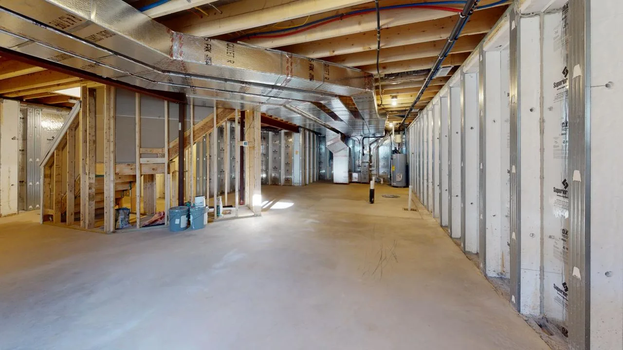 Spacious unfinished basement with exposed wooden beams, metal ducts, and studded walls. Concrete floor is clear and open, with construction materials and tools visible near wooden stairs on the left.