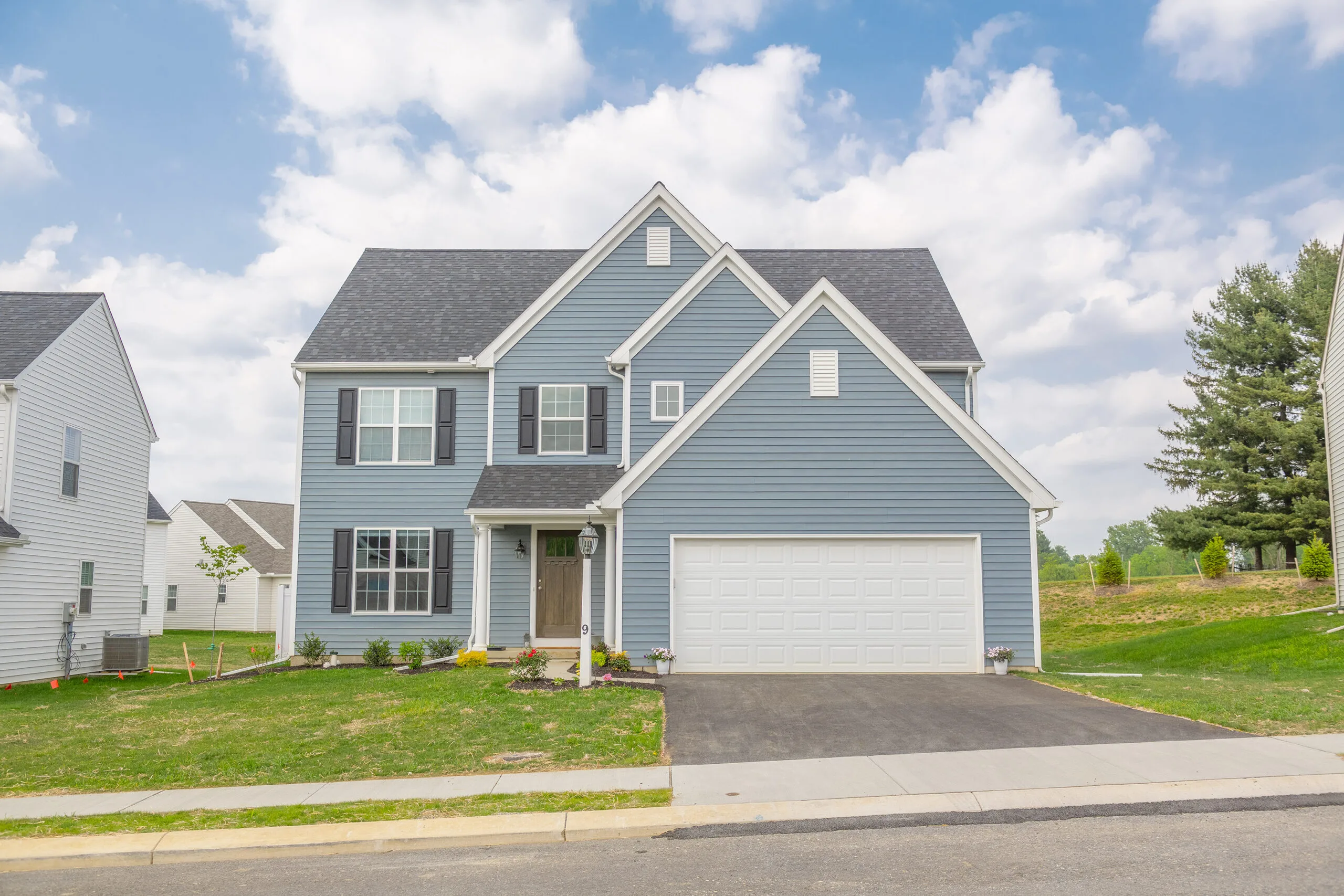 A modern two-story blue house with white trim, a gray shingle roof, and a double garage, set on a green lawn under a partly cloudy sky.