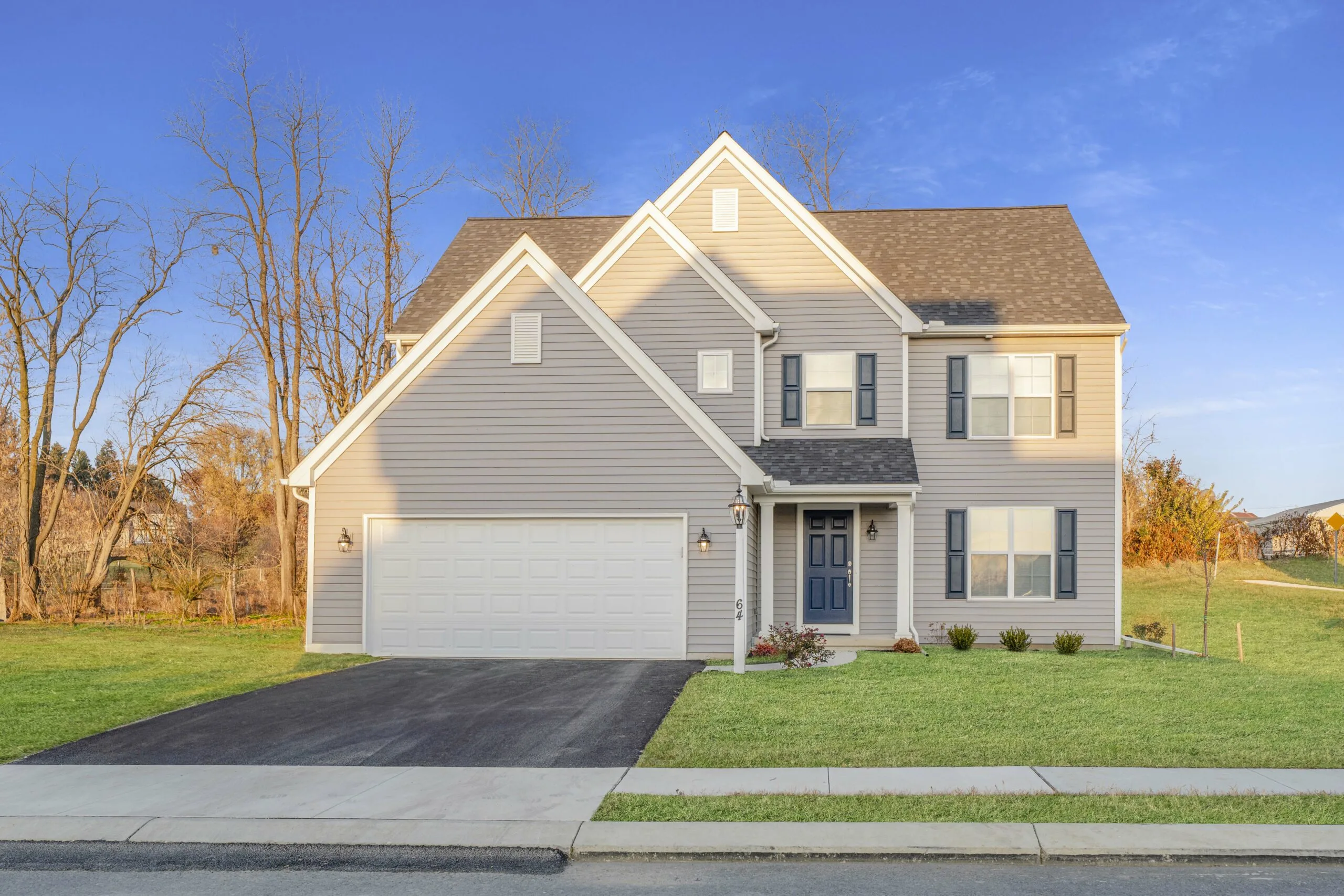 A two-story beige house with a double garage, blue front door, and well-kept lawn, set against a clear blue sky and bare trees in the background.