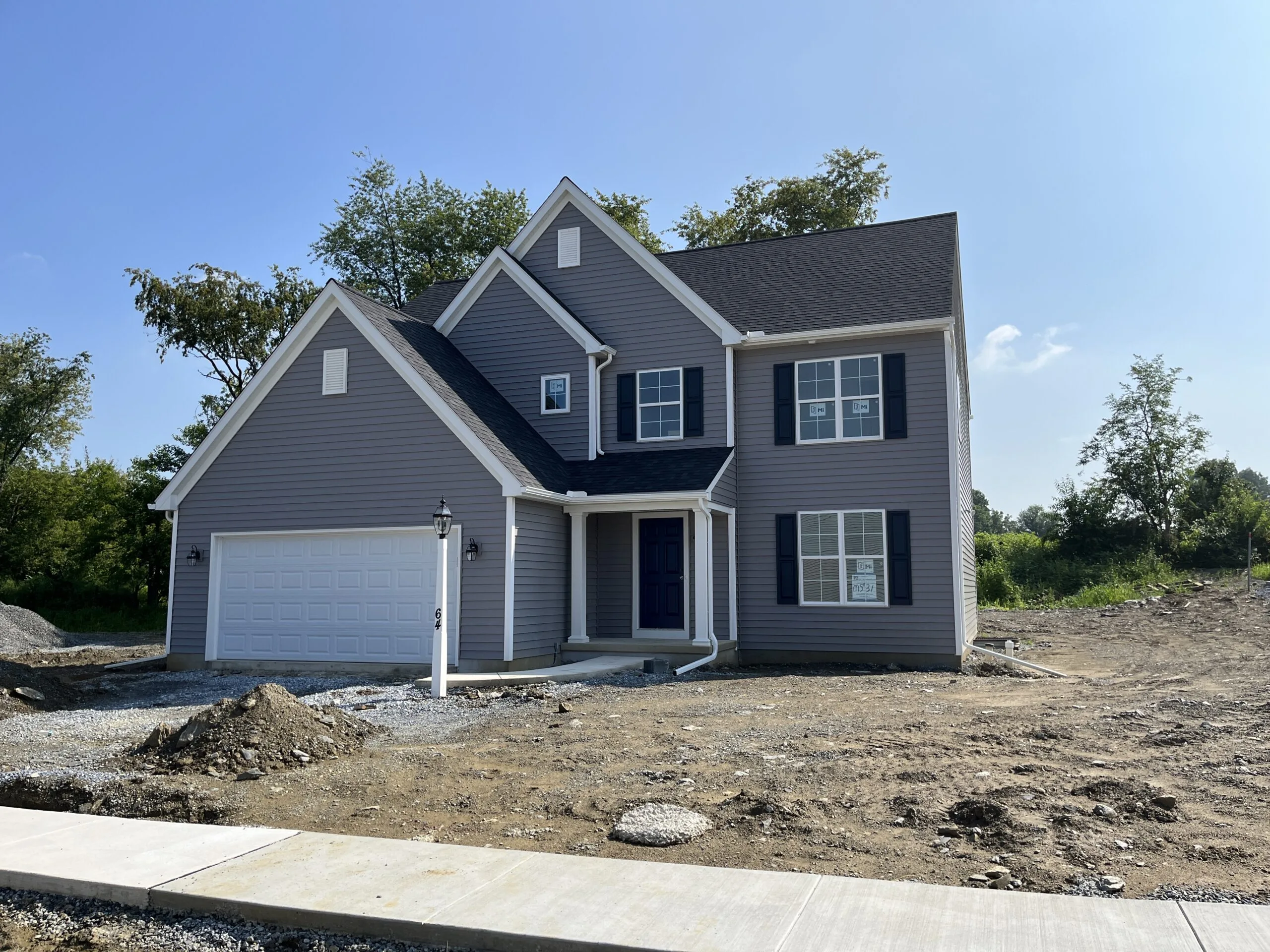 A newly built two-story gray house with white trim and a double garage, standing on a dirt lot with no landscaping, under a clear blue sky.