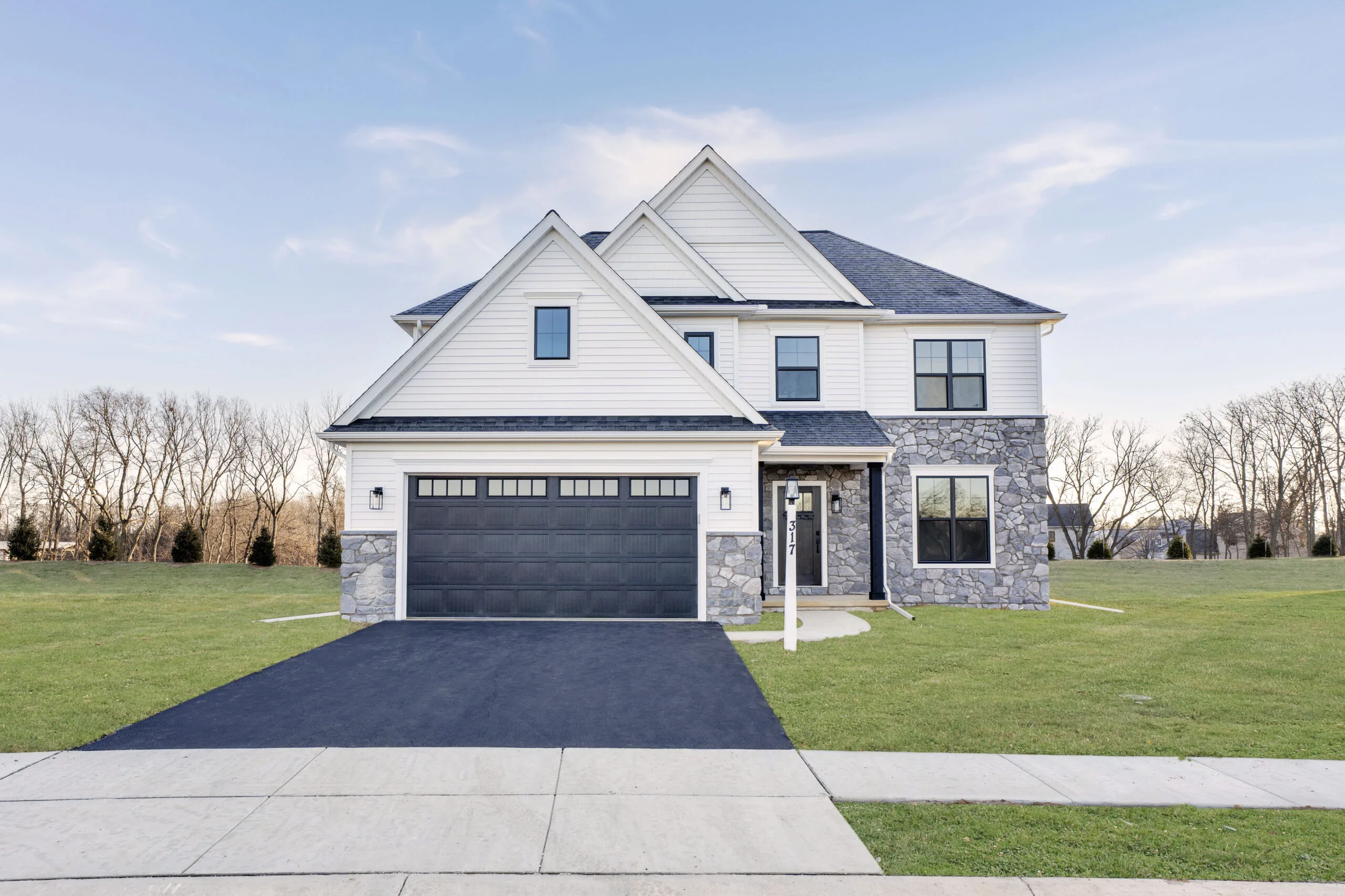 A modern two-story house with light siding, stone accents, a black double garage door, large windows, and a paved driveway, set on a grassy lawn with trees and a clear sky in the background.