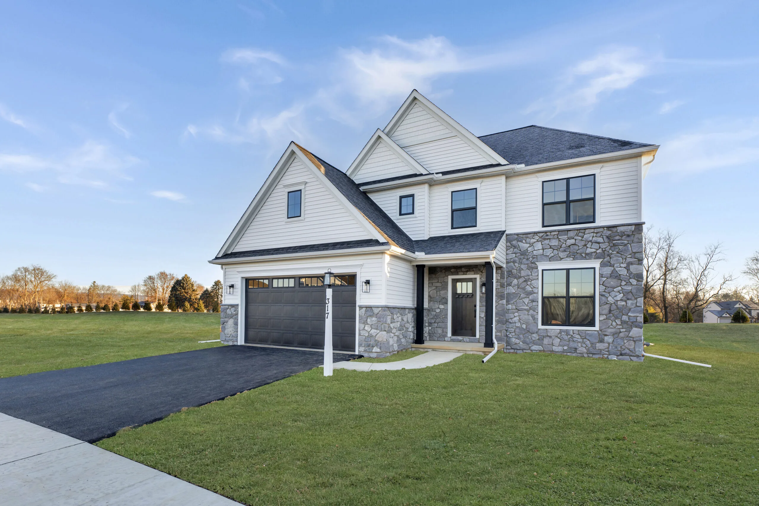 A modern two-story house with white siding and stone accents, attached two-car garage, and large green lawn under a clear blue sky.
