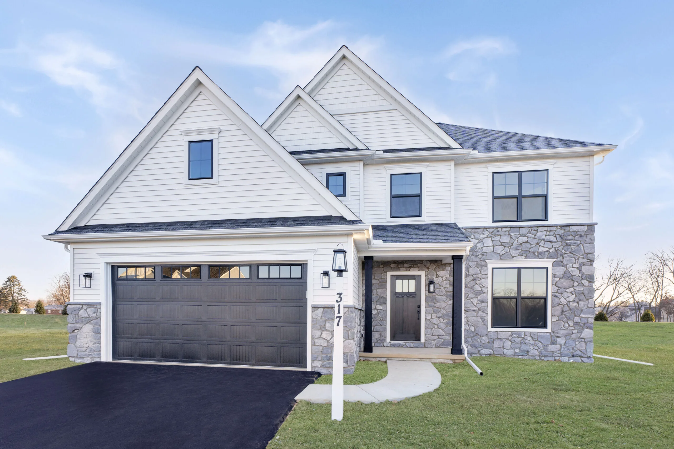 Modern two-story house with light siding and stone accents, large black garage door, front porch, black front door, lamppost with house number 317, and manicured lawn under a clear blue sky.