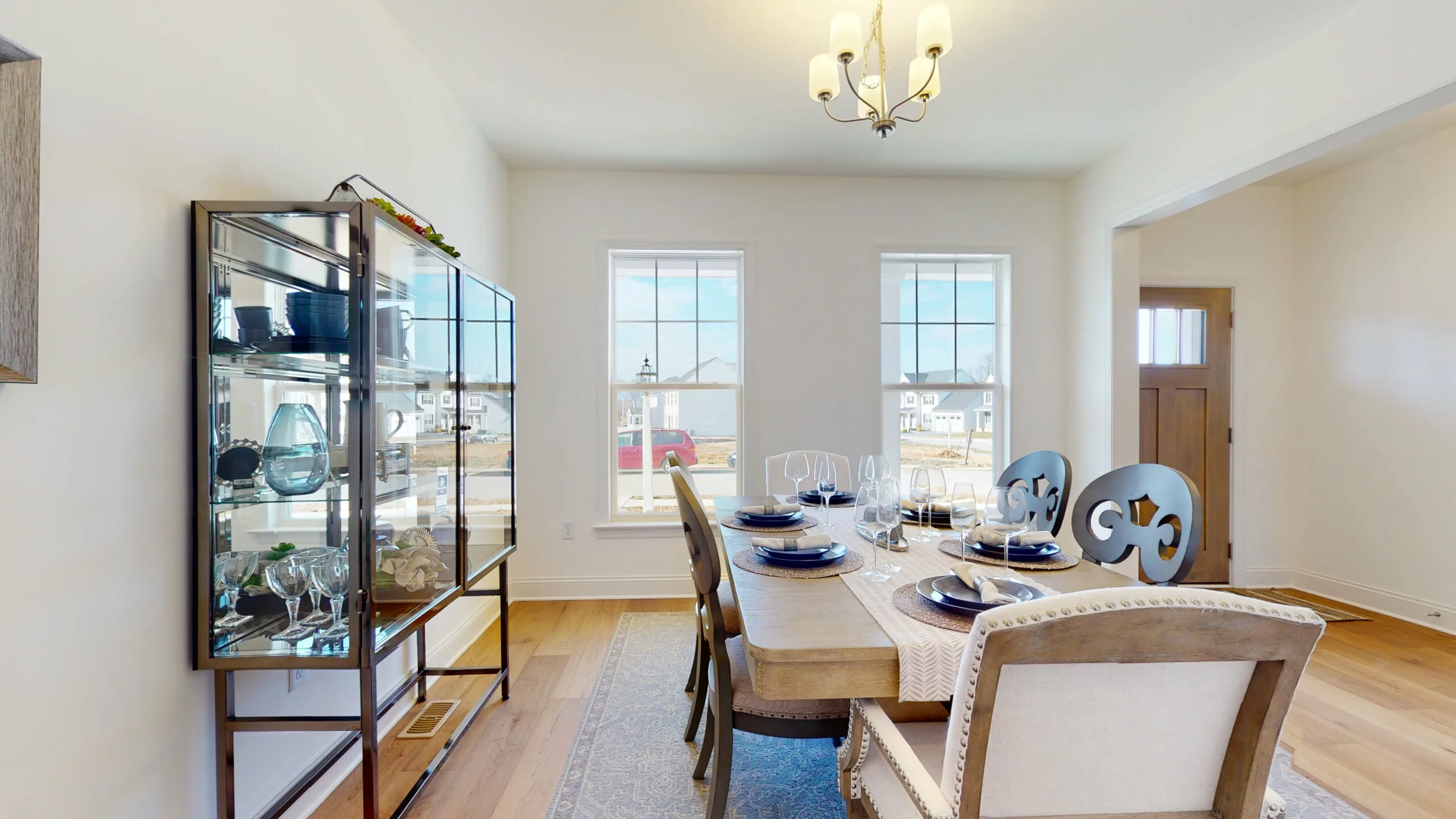Bright dining room with a wooden table set for six, blue and white tableware, glass-front cabinet with decor items, large windows letting in natural light, and a chandelier overhead.