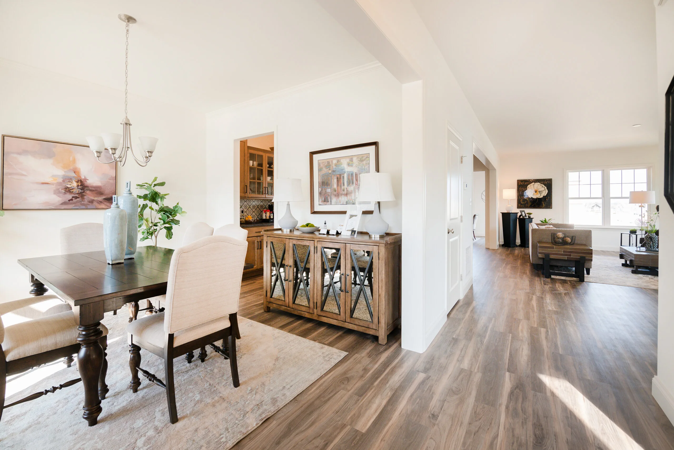 Bright, modern dining area with a wooden table and upholstered chairs on a beige rug, next to a hallway with wood flooring leading to a spacious living room with large windows and neutral decor.