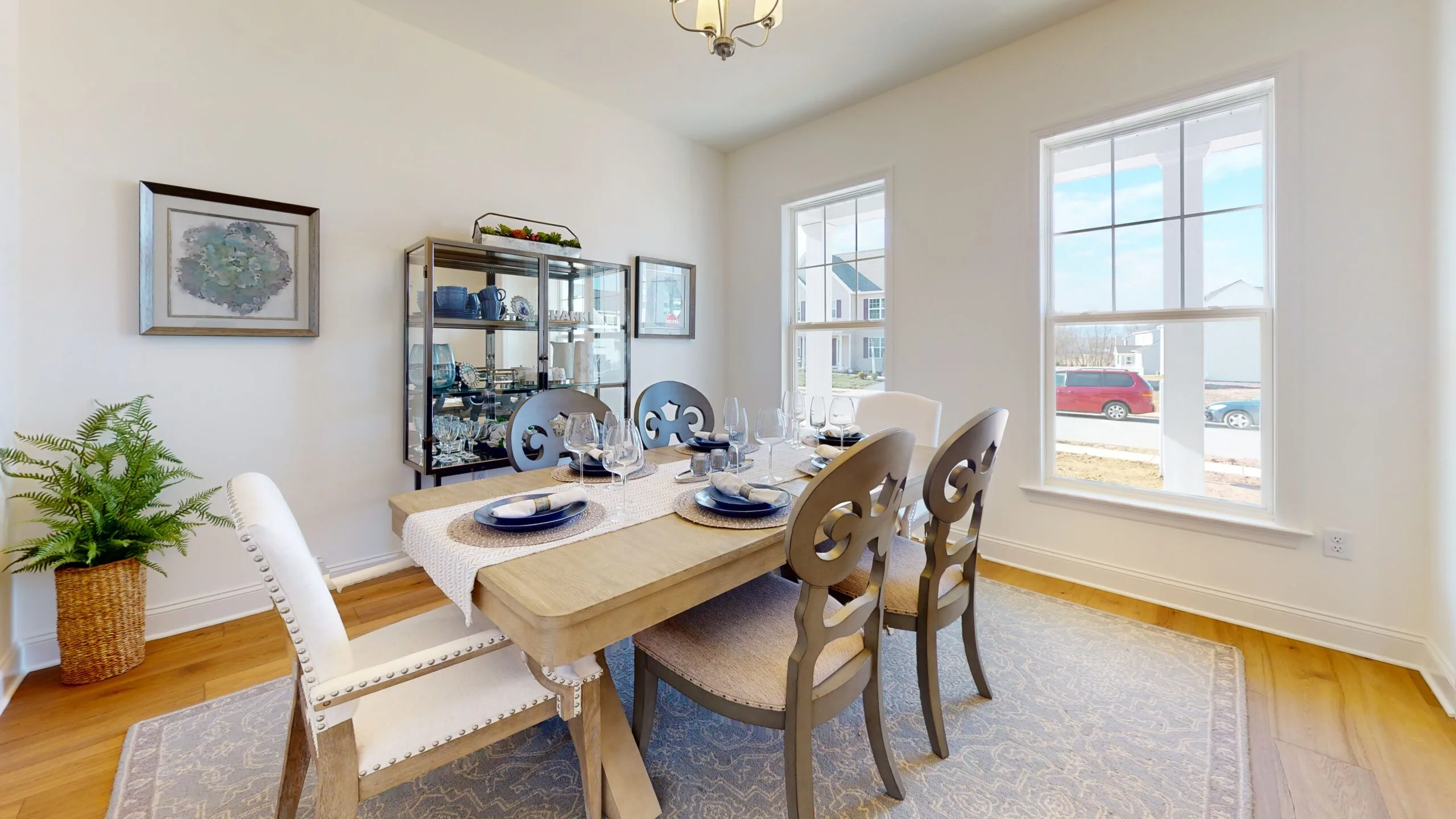 A bright dining room with a wooden table set for six, featuring light upholstered chairs, a china cabinet with dishes, two large windows letting in natural light, and a plant in the corner.