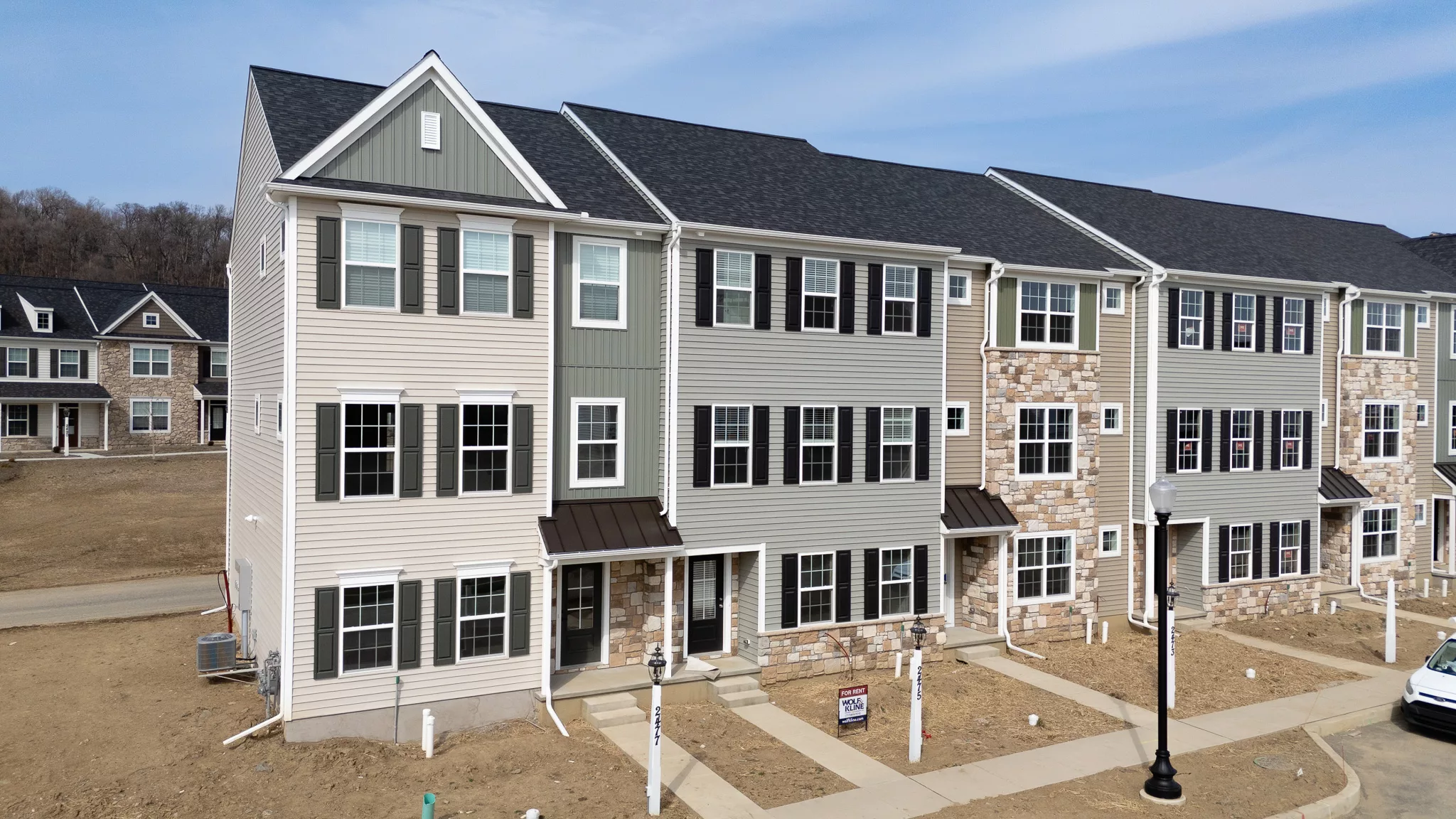 A row of new, multi-story townhouses with beige siding and brick accents, large windows, and small covered entryways. The yards are bare dirt, indicating recent construction. A For Sale sign is visible near one entrance.