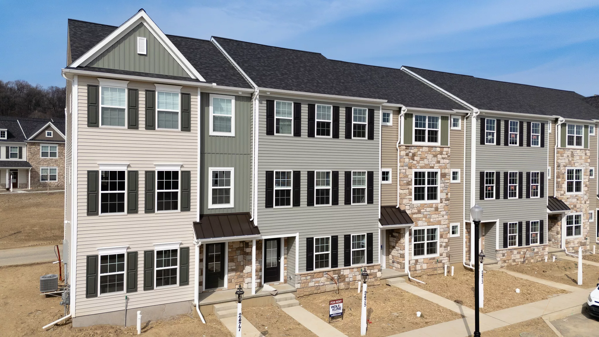 A row of newly-built three-story townhouses with beige and gray siding, multiple large windows, and stone accents. The area in front is bare dirt, with sidewalks and a “For Sale” sign visible.