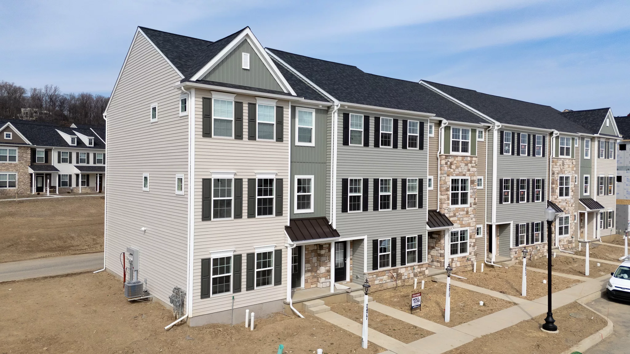 A row of newly built, three-story townhouses with beige siding and stone accents, set on a bare dirt lot with minimal landscaping under a partly cloudy sky.