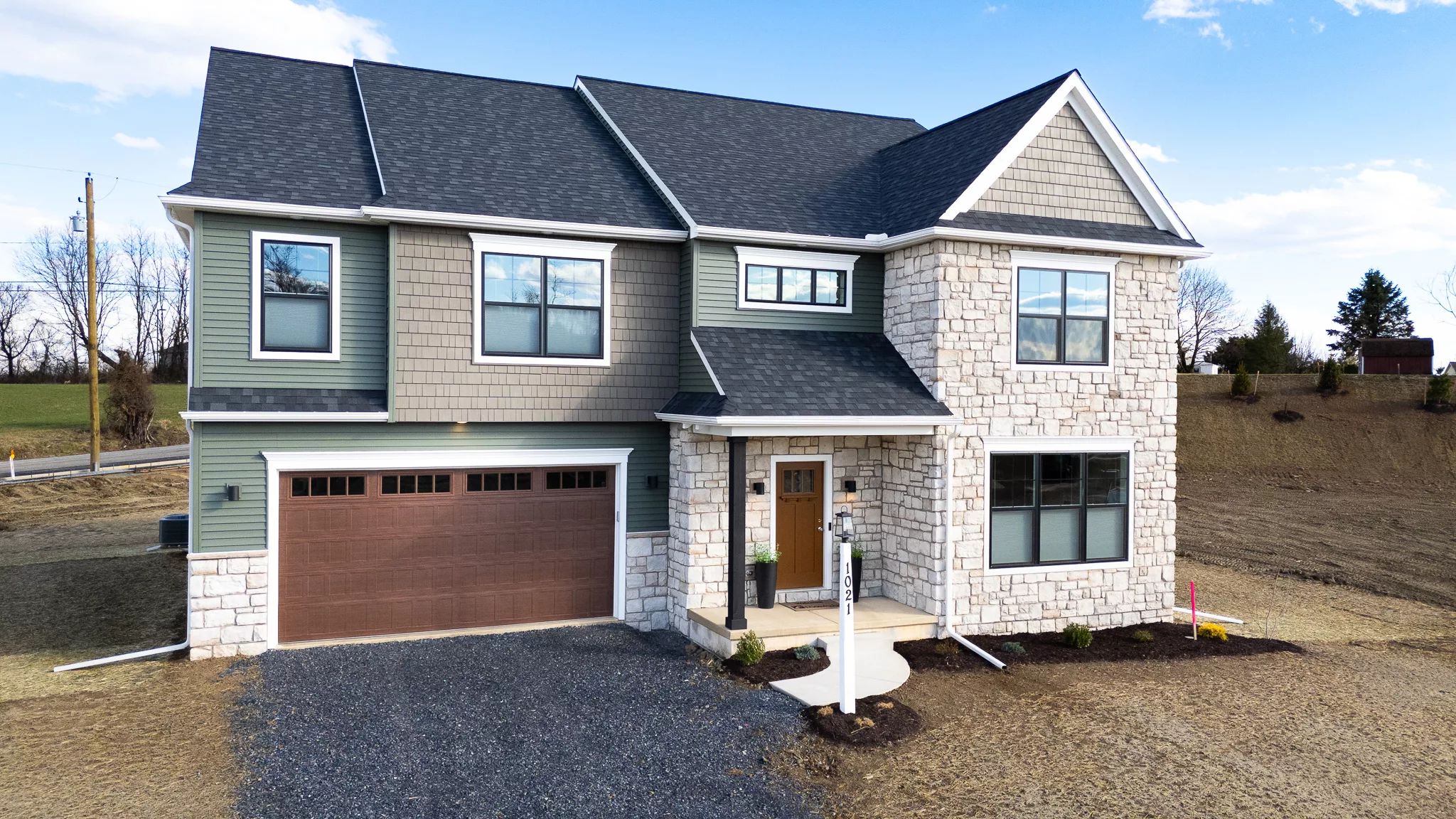 Two-story modern house with a stone and siding exterior, large windows, and a brown double garage. The house sits on a bare, unfinished yard with minimal landscaping and a gravel driveway.