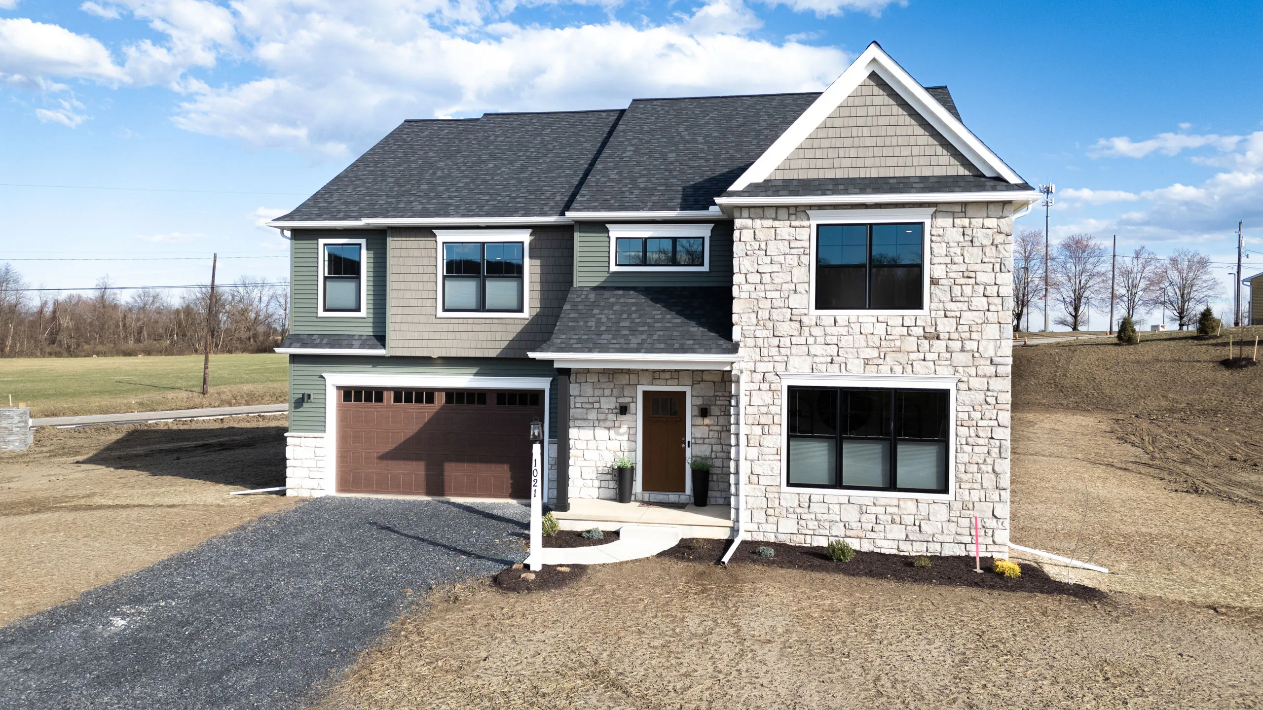 Two-story modern suburban house with mixed stone and siding exterior, large front windows, and a two-car garage, surrounded by a bare, brown yard under a partly cloudy blue sky.