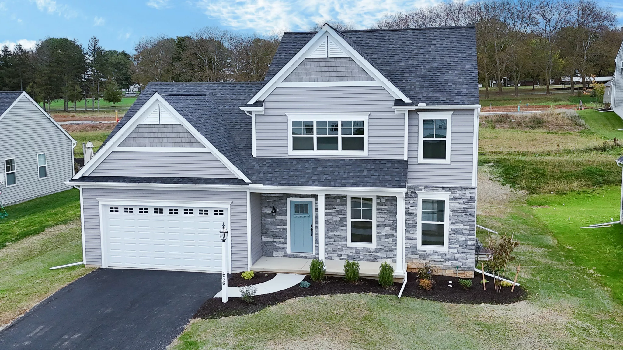 A two-story, modern suburban house with gray siding, stone accents, a double garage, black-shingled roof, and a front porch. The home sits on a neatly landscaped yard with grass and small shrubs.