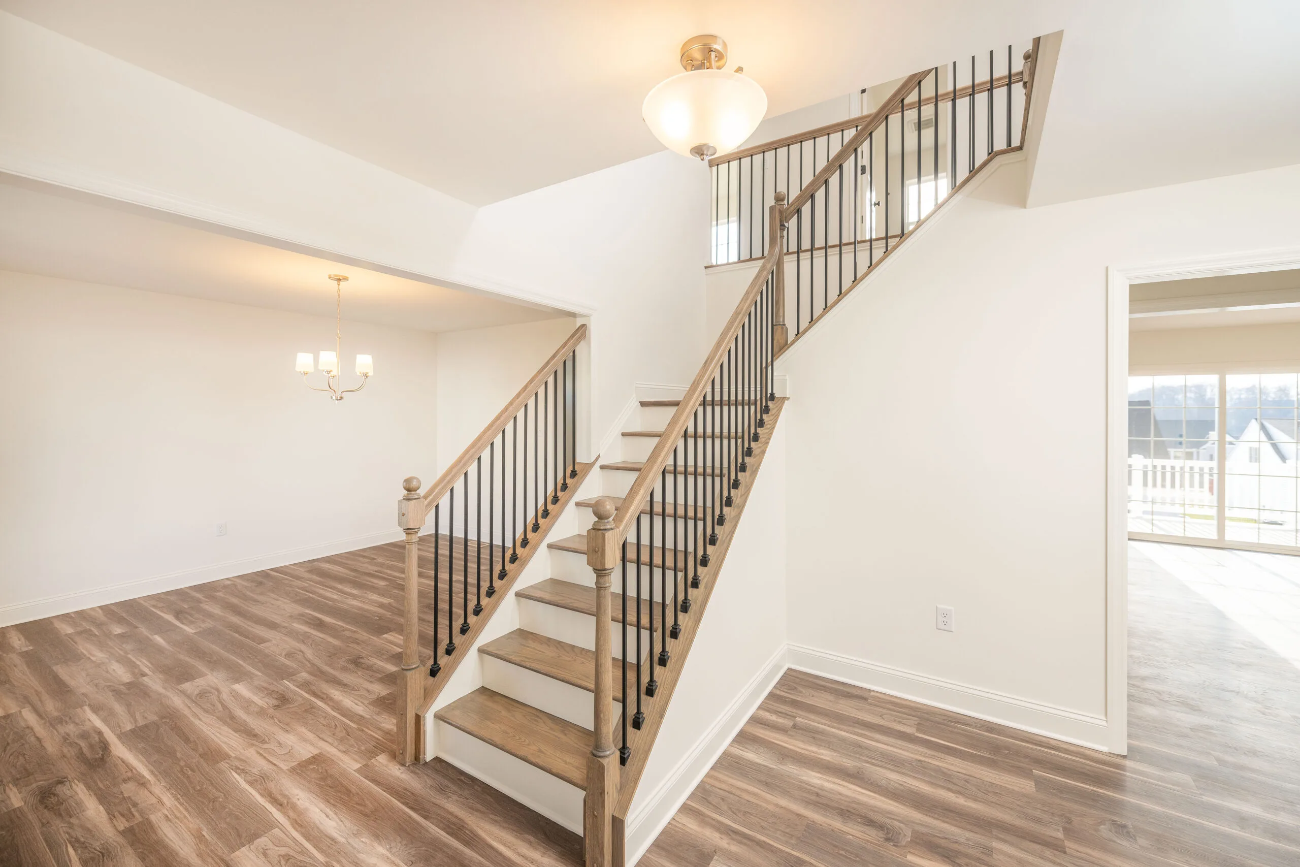 A bright, modern home interior features a wooden staircase with black metal railings, light wood flooring, white walls, and two ceiling light fixtures in open adjacent rooms. Large windows let in natural light.