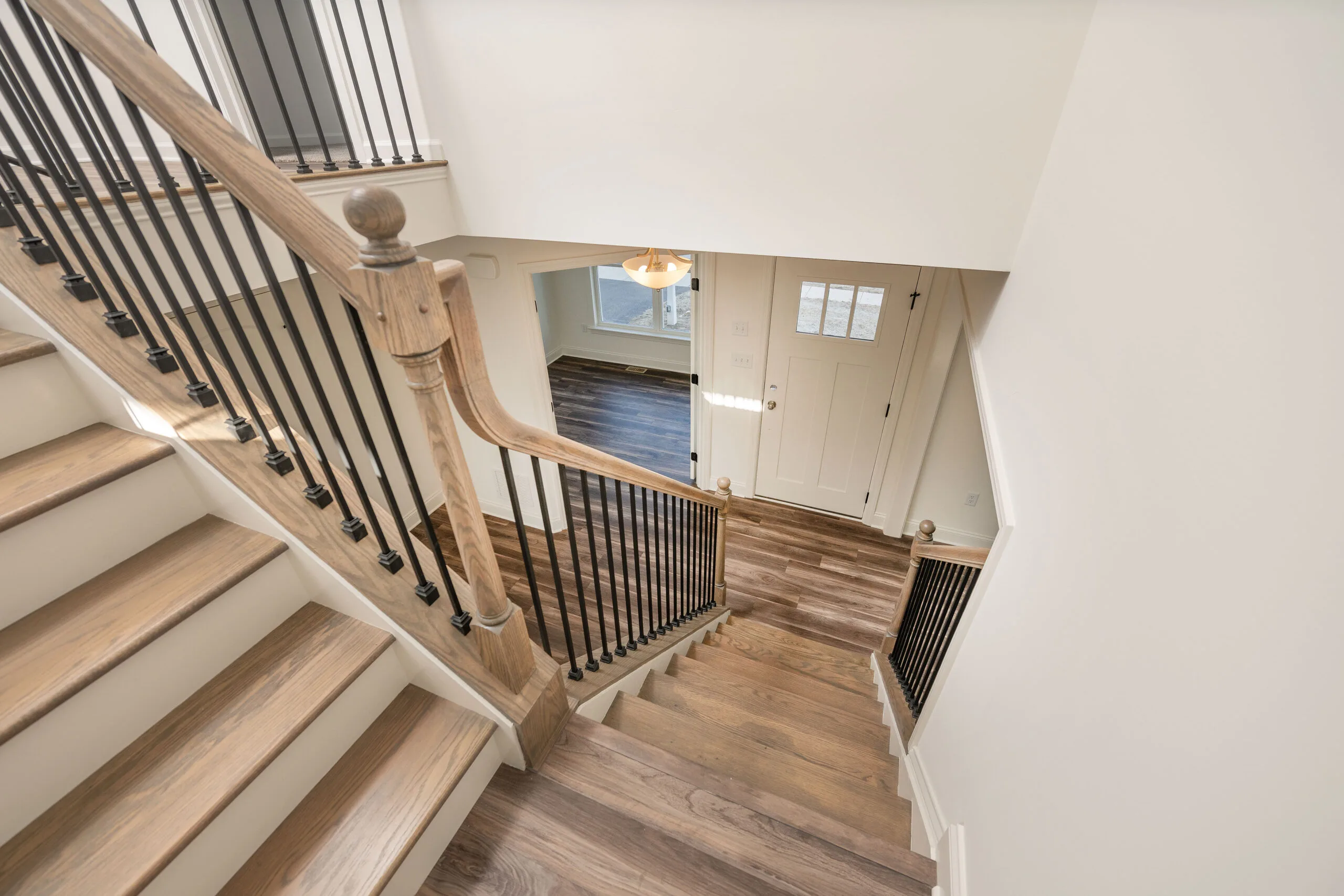 View from the top of a wooden staircase with black metal balusters, leading down to a small foyer with a white front door, sidelight window, and wood-patterned flooring. The space is bright and airy.