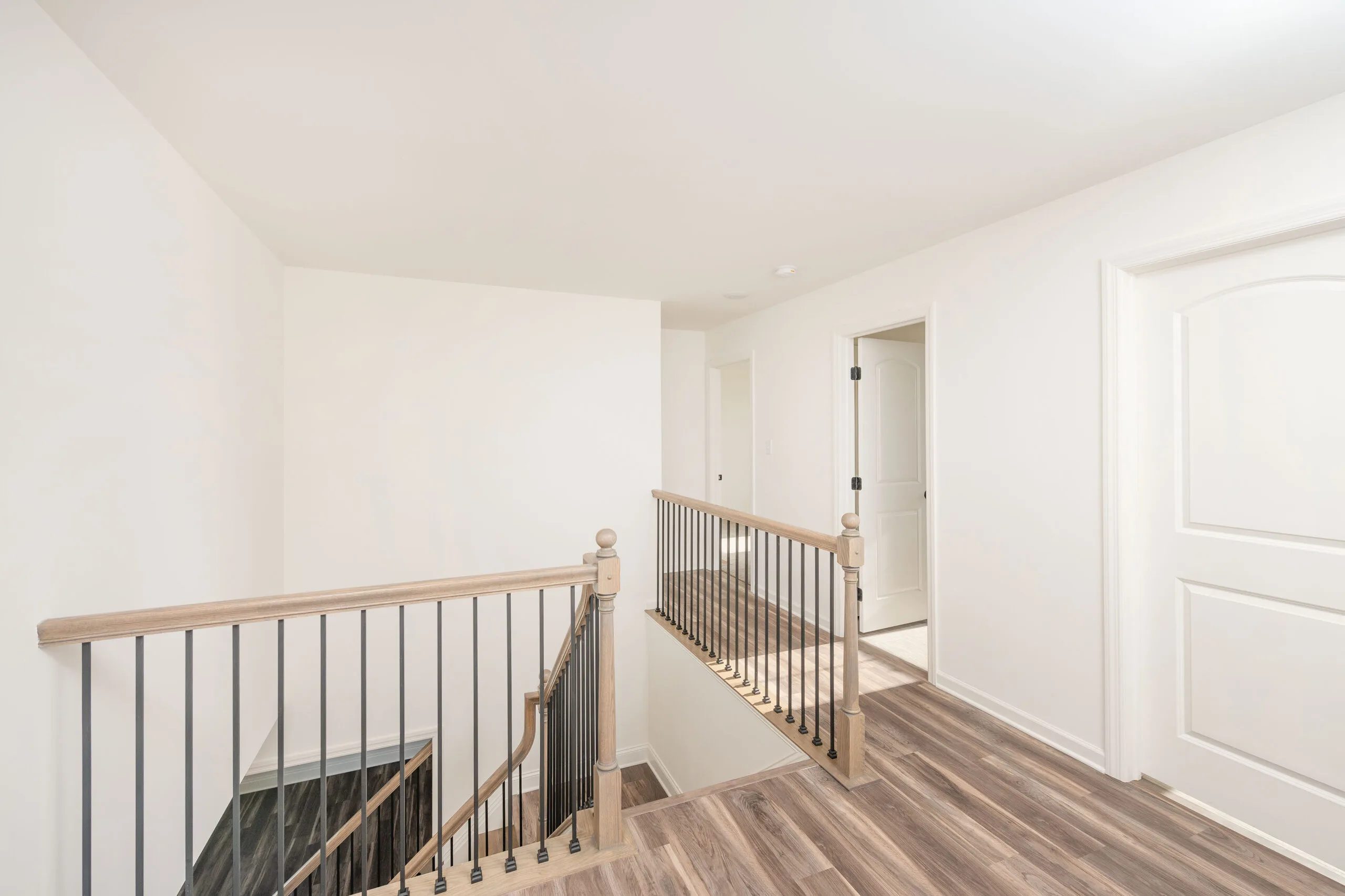 A bright, modern hallway with light wood flooring, white walls, a staircase with wooden handrails and black spindles, and several white doors leading to adjacent rooms.