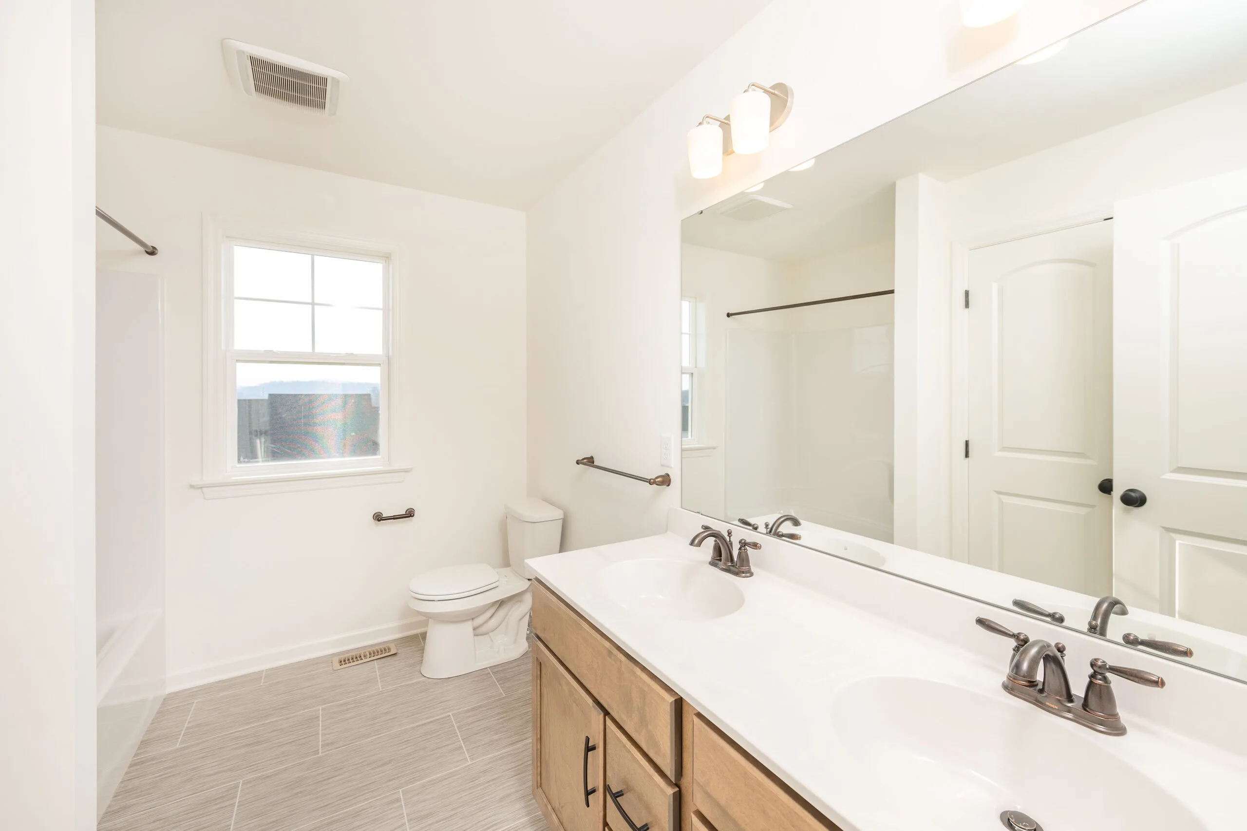 Bright, modern bathroom with light tile floor, double sink vanity with wood cabinets, large mirror, toilet, bathtub with shower curtain rod, and a window letting in natural light.