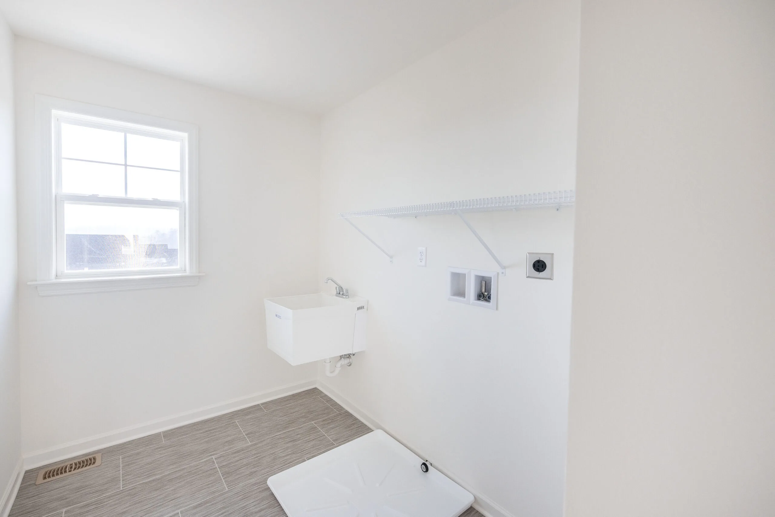 A bright, empty laundry room with beige tiled floor, white walls, a utility sink, overhead wire shelf, washer and dryer hookups, and a window letting in natural light.