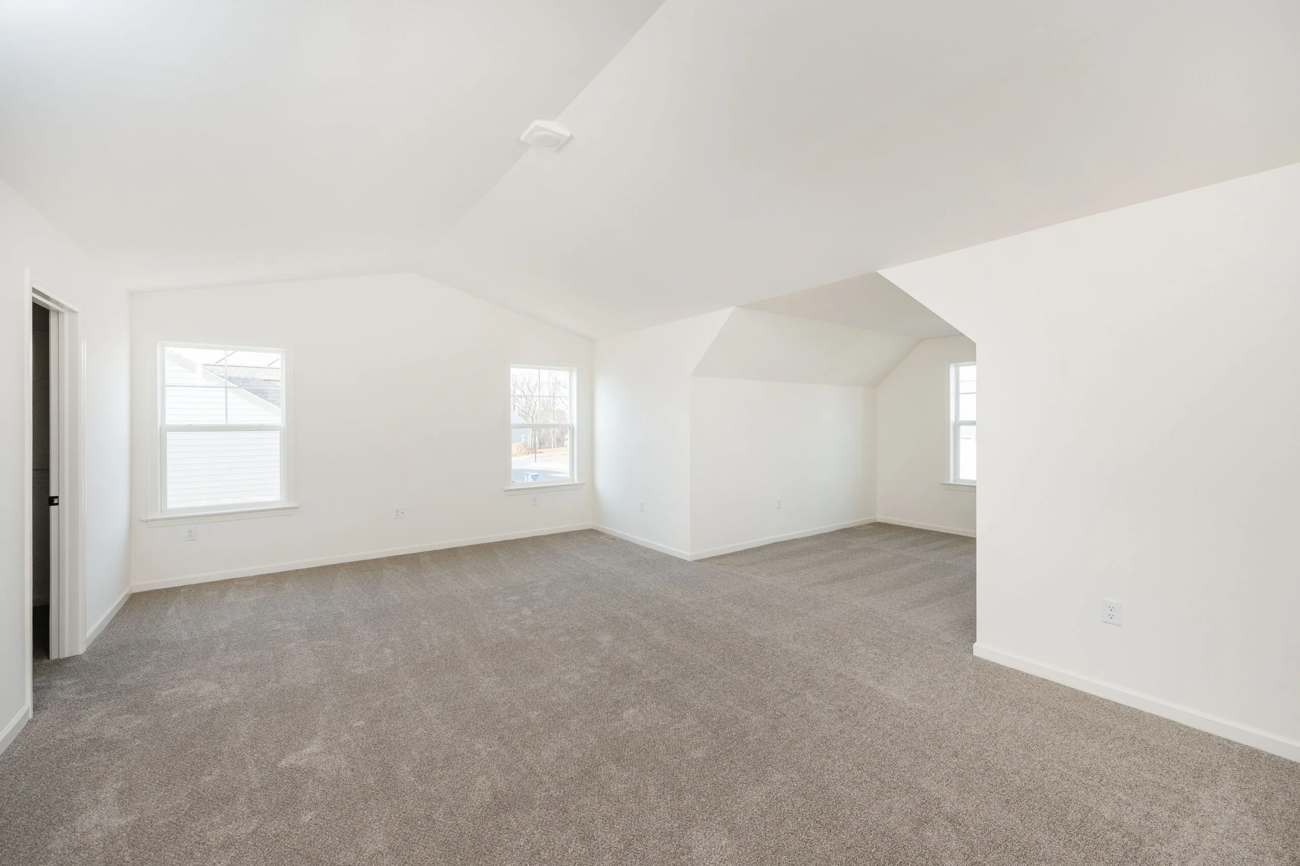 Spacious, empty room with light beige carpet and white walls, two large windows letting in natural light, and a doorway on the left side. The ceiling is angled and the space appears clean and bright.