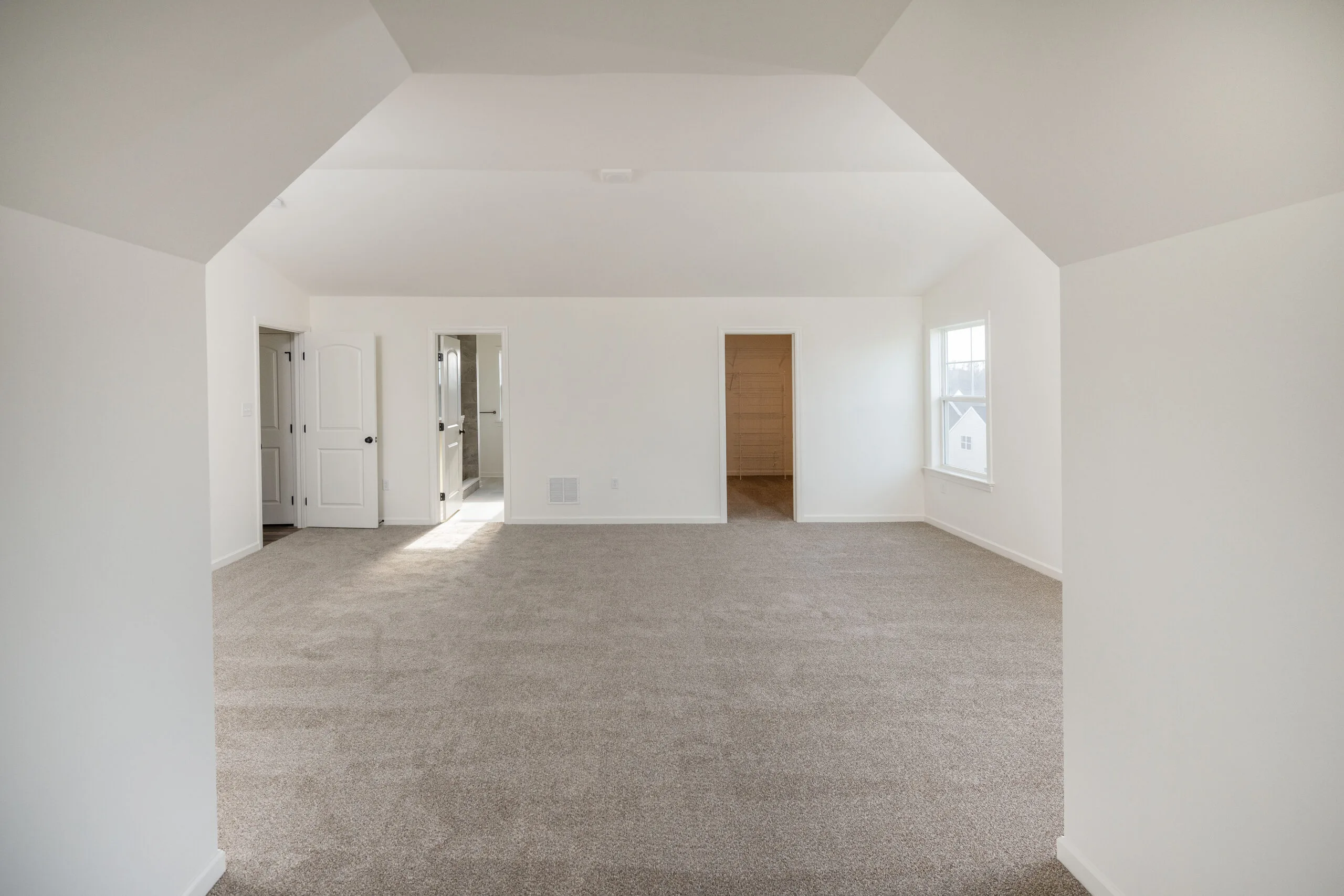 A spacious, empty room with light beige carpet, white walls, and a sloped ceiling. There are two open doorways and a window letting in natural light on the right side.