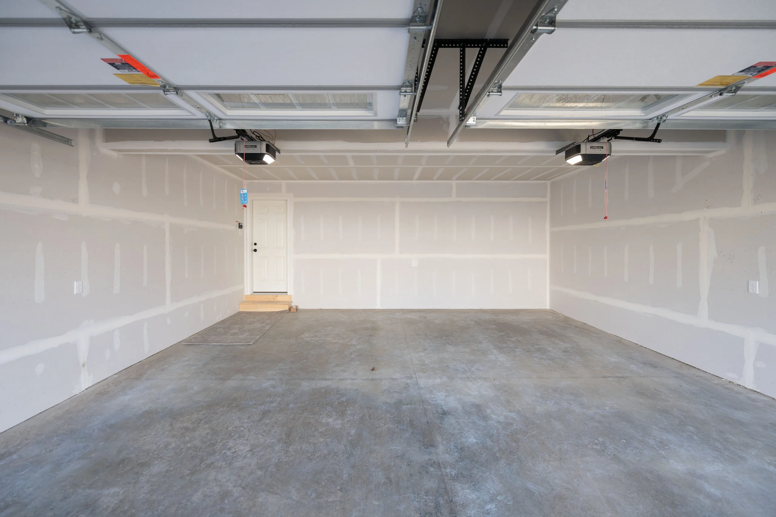 Empty two-car garage with unfinished drywall walls, concrete floor, and two open garage doors. A white door with a small step leads to the interior of the house. Ceiling-mounted garage door openers are visible.