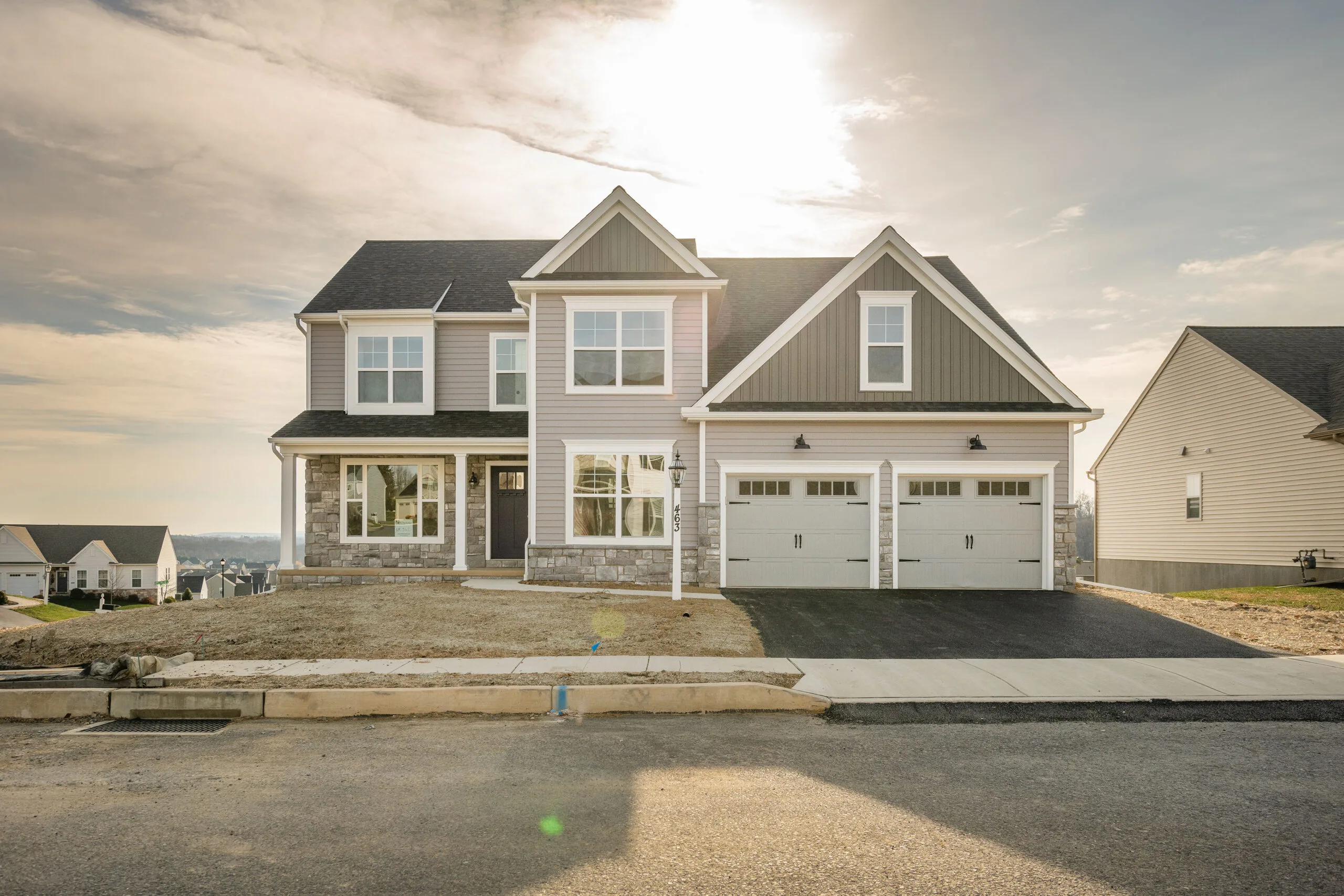 Two-story modern suburban house with a double garage, light-colored siding, and stone accents. The driveway is freshly paved, and the front yard is unfinished. The sky is partly cloudy with the sun shining above the home.