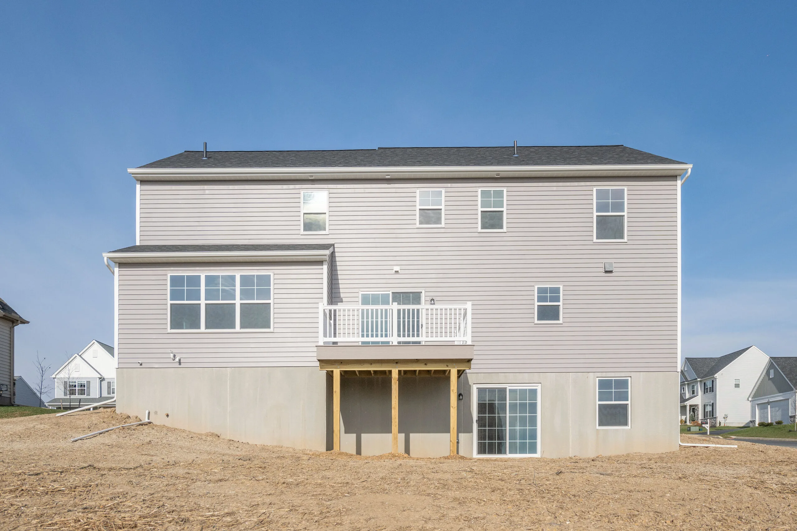 A newly constructed two-story house with light gray siding, white trim, and a small balcony. The backyard is unfinished with bare dirt, and there are large windows and a sliding glass door on the lower level.