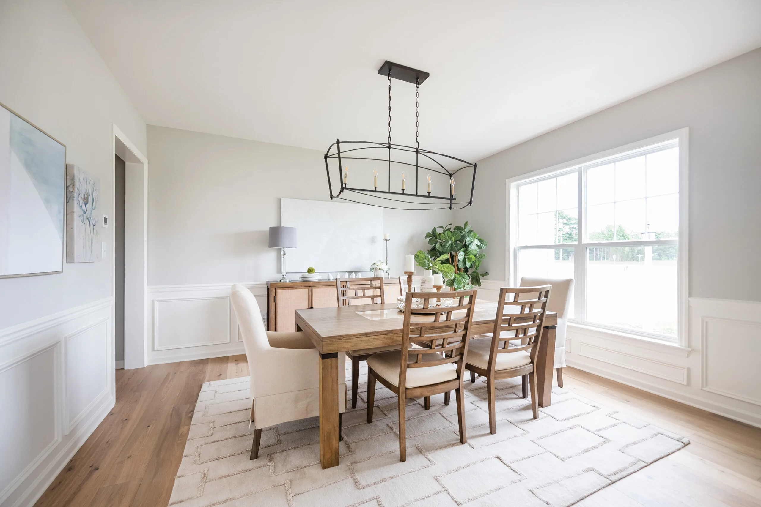 A bright, modern dining room with a wooden table, six chairs, a cushioned bench, a large window letting in natural light, a geometric chandelier, a sideboard, and a potted plant in the corner.