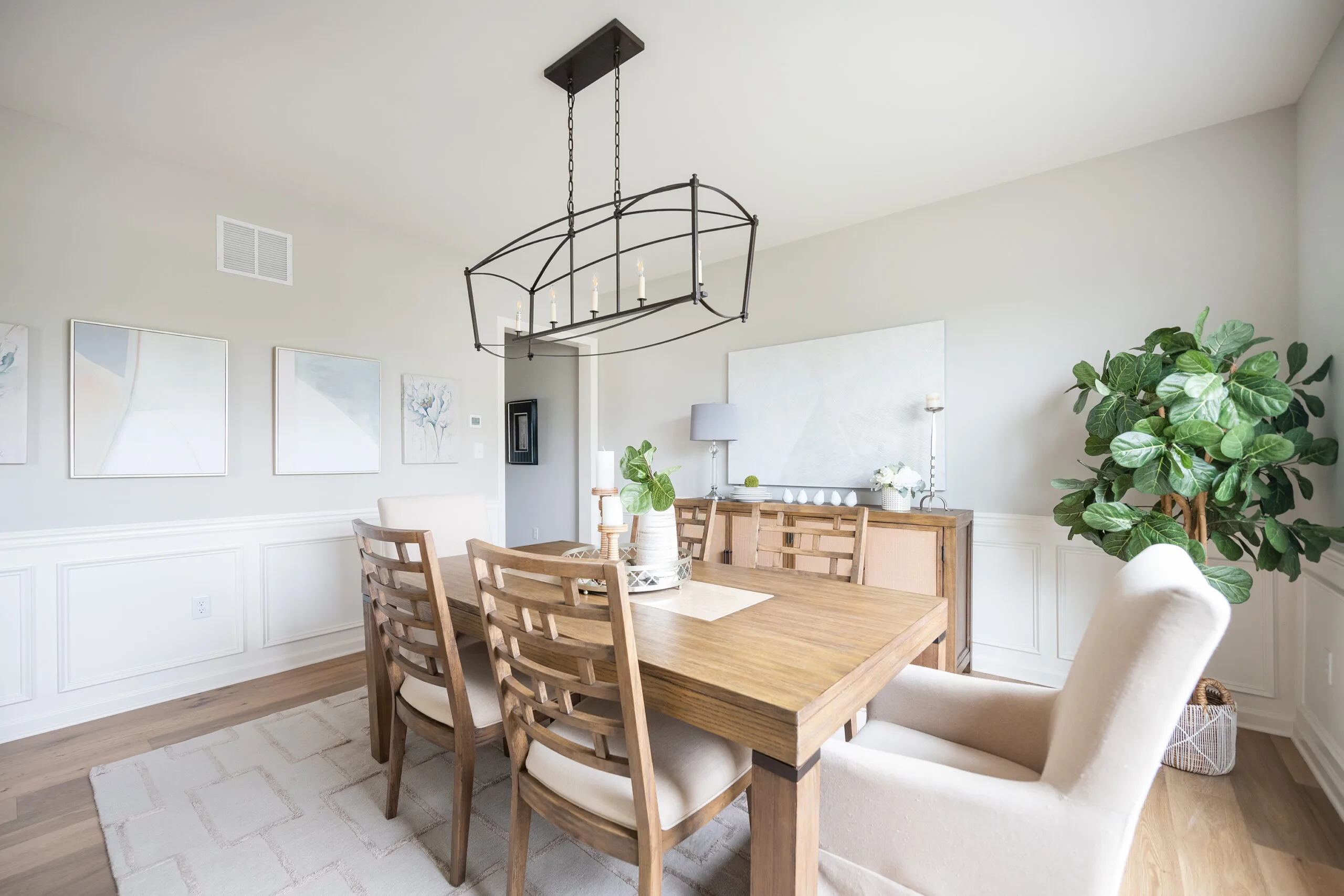 Bright dining room with a wooden table, six beige chairs, a modern black chandelier, wall art, a potted plant, and large windows letting in natural light. Decor is minimalist with neutral tones.