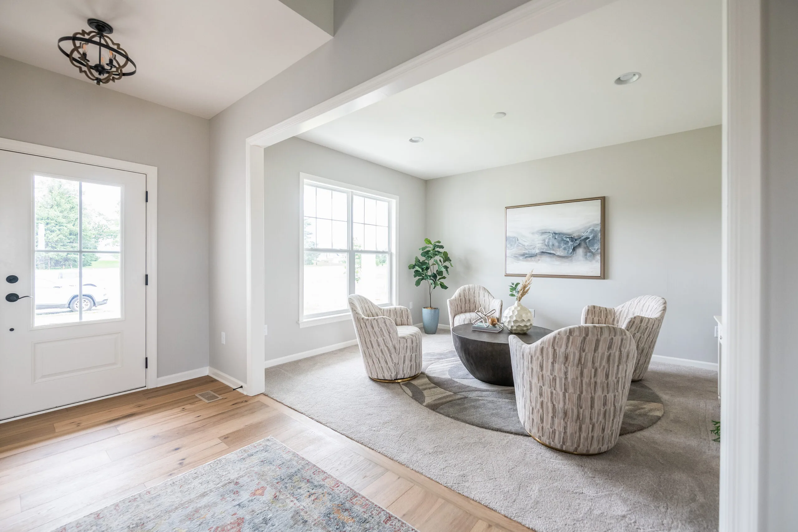 Bright, modern dining room with light gray walls, large window, round table, five patterned chairs, abstract wall art, potted plant, and wood flooring transitioning to carpet. Door and rug visible in the foreground.