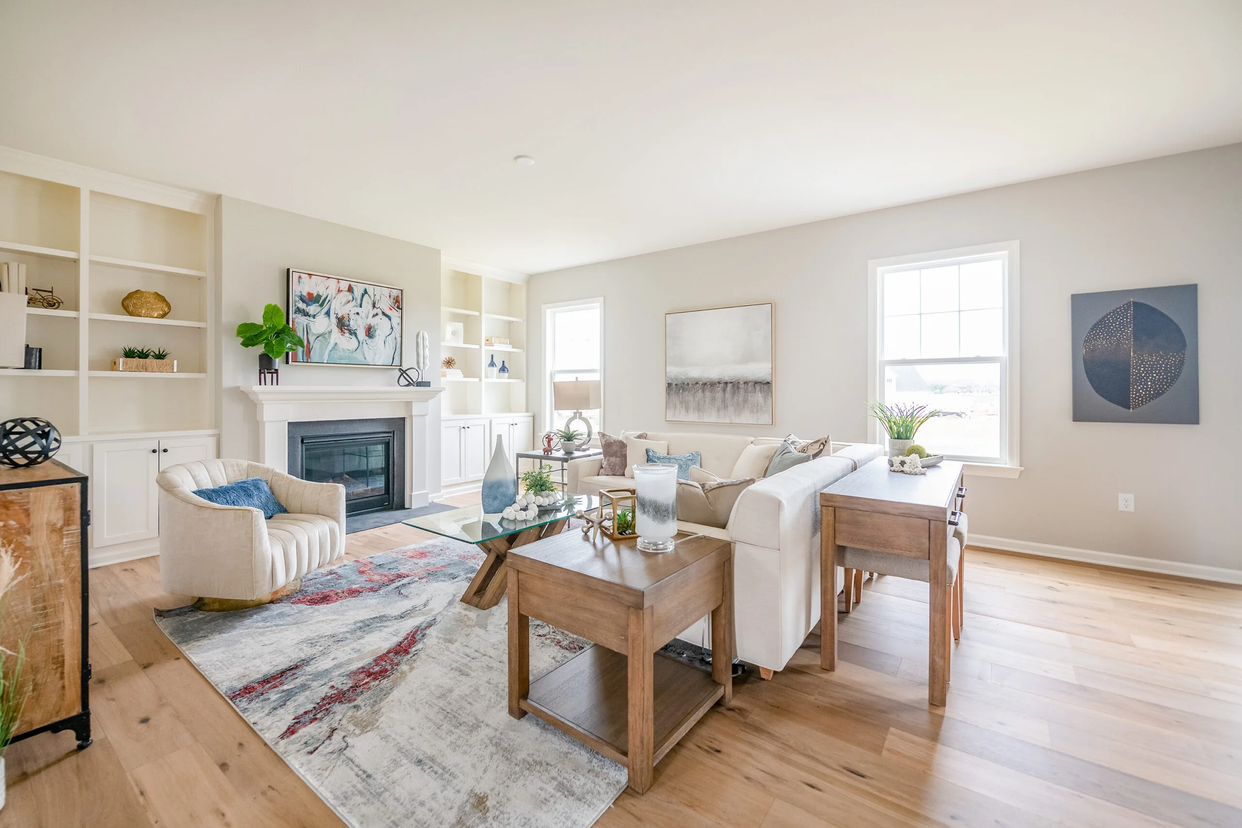 Bright living room with light wood floors, cream walls, a white fireplace, modern furniture, abstract art, shelves with decor, large windows, and indoor plants. The space feels airy and welcoming.