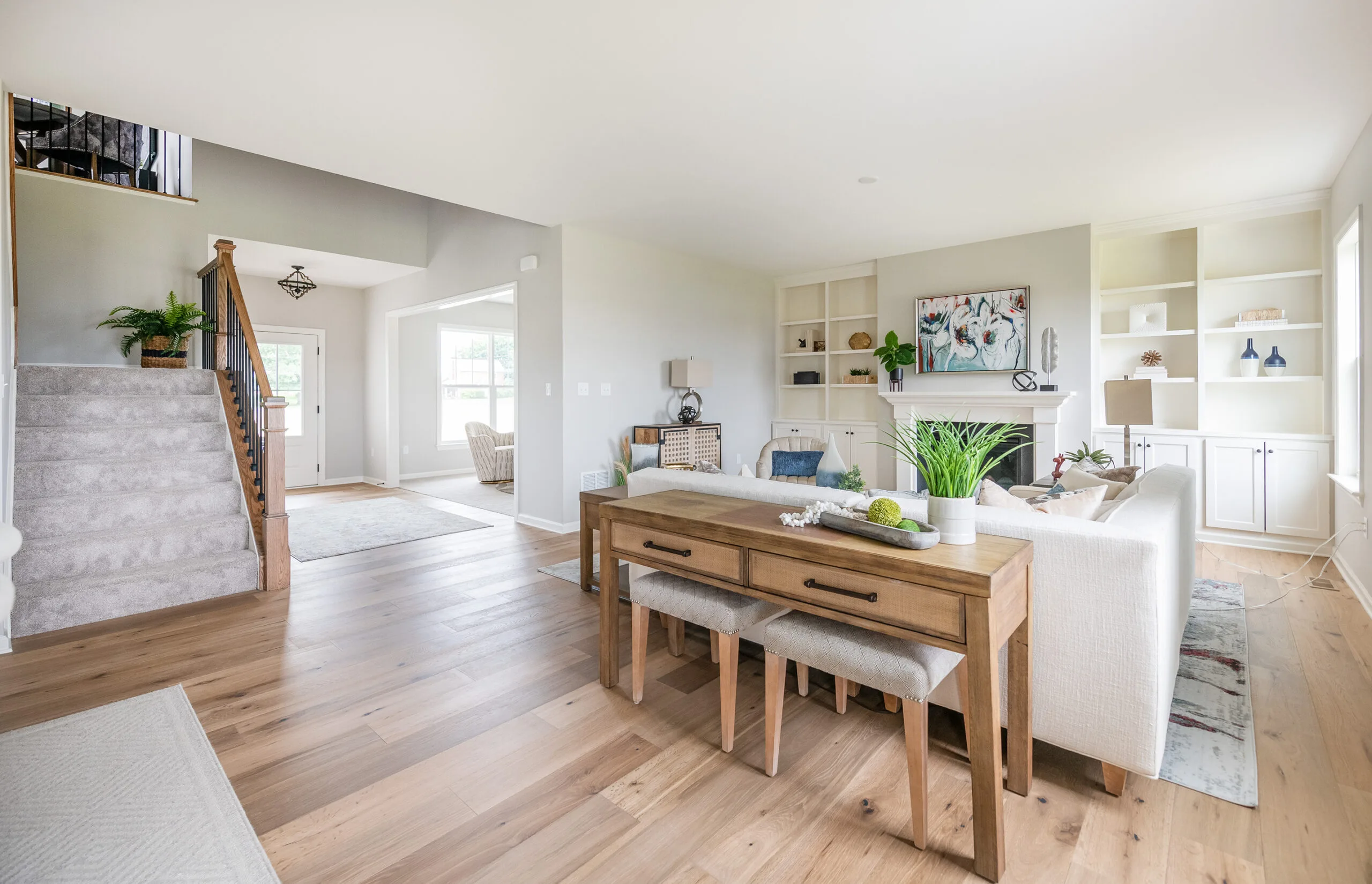 Bright, modern living and dining area with light wood floors, a white sofa, wooden dining table, built-in shelves, large windows, and a staircase with beige carpet on the left. Green plants add a touch of color.