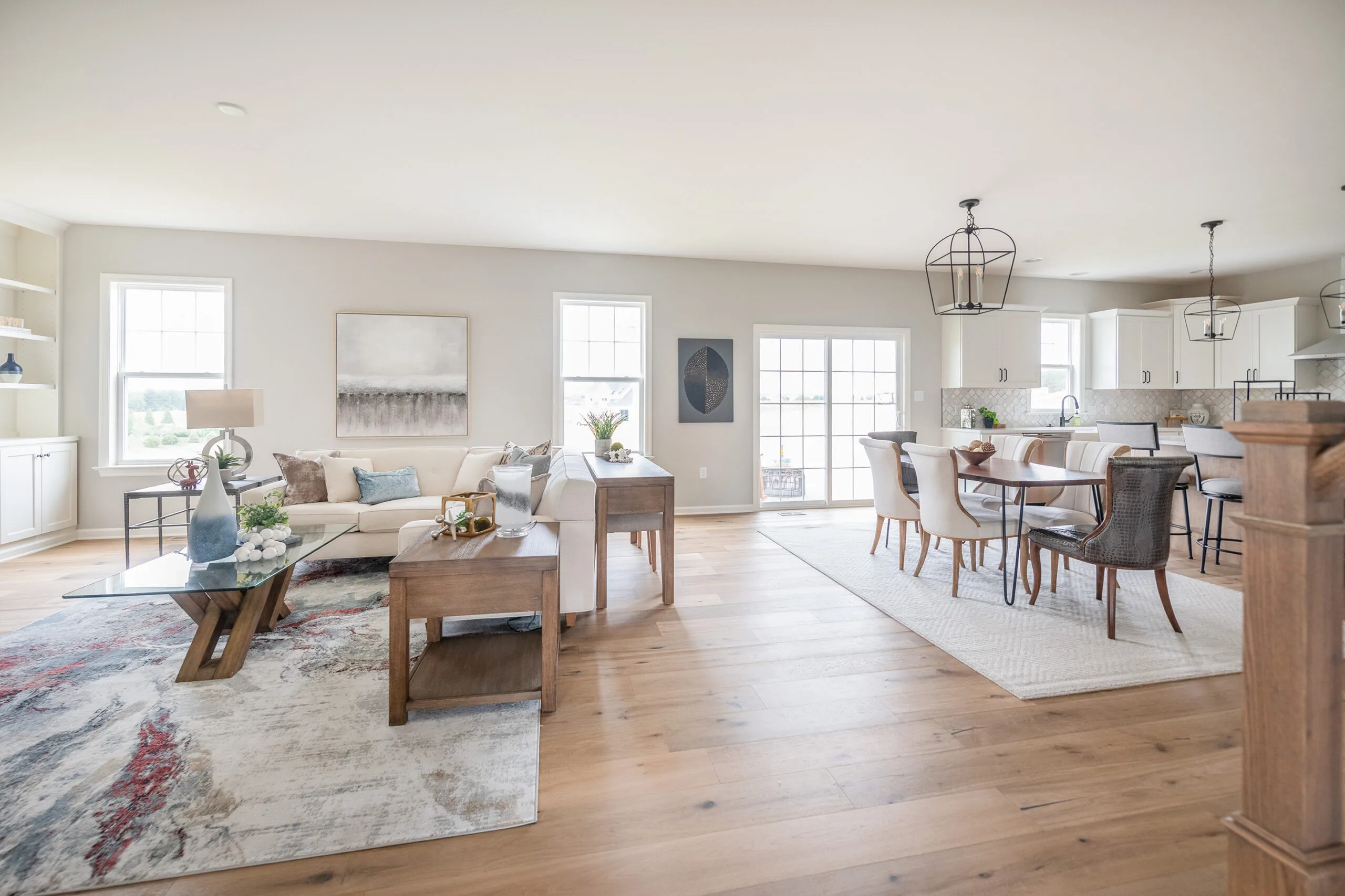 Bright open-concept living and dining area with hardwood floors, modern furniture, neutral colors, large windows, and contemporary decor; the kitchen is visible in the background.