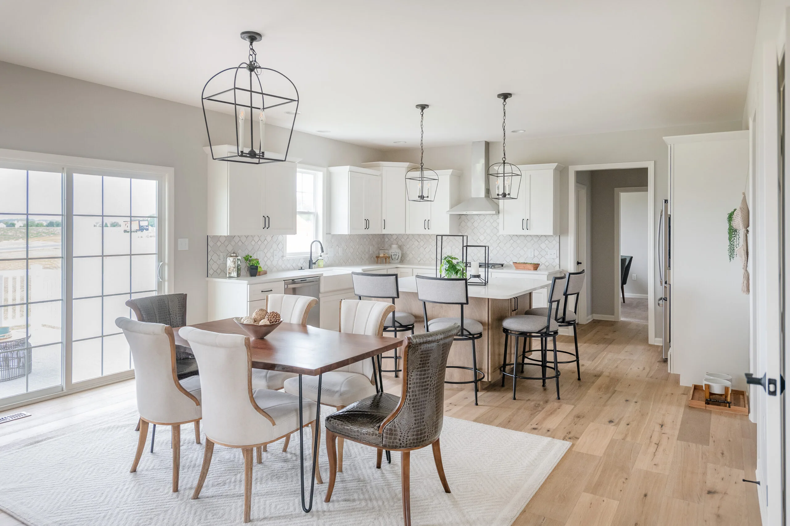 A bright, modern kitchen and dining area with white cabinets, light wood floors, a wooden dining table with mixed chairs, a kitchen island with stools, and large windows letting in natural light.