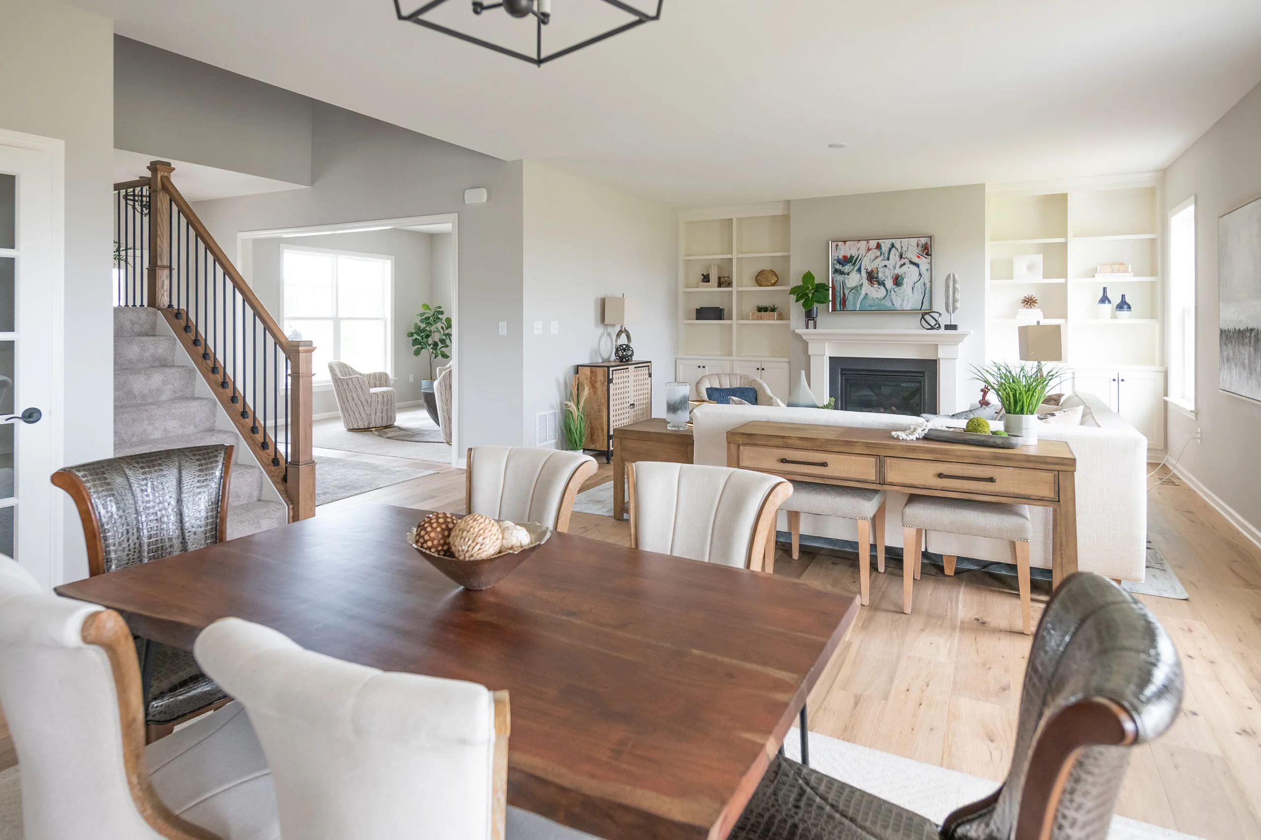 Open-plan living and dining area with a wooden table and upholstered chairs in the foreground, a cozy sitting area with sofas, shelves, and a fireplace in the background, and stairs leading upstairs.