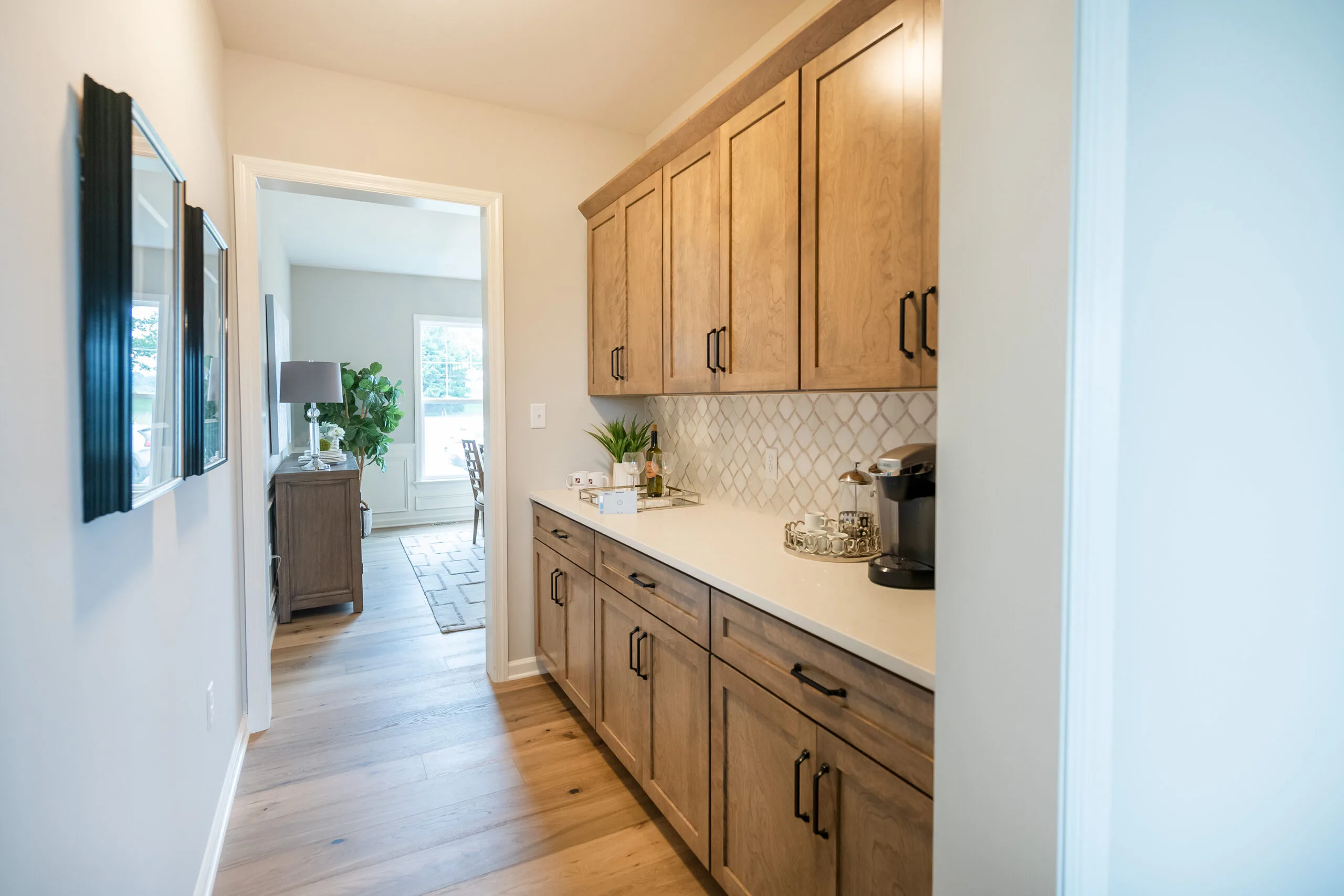 A narrow hallway with light wooden flooring, beige cabinets with black handles, a white countertop, and a coffee maker. The hallway leads to a bright room with windows and a side table.
