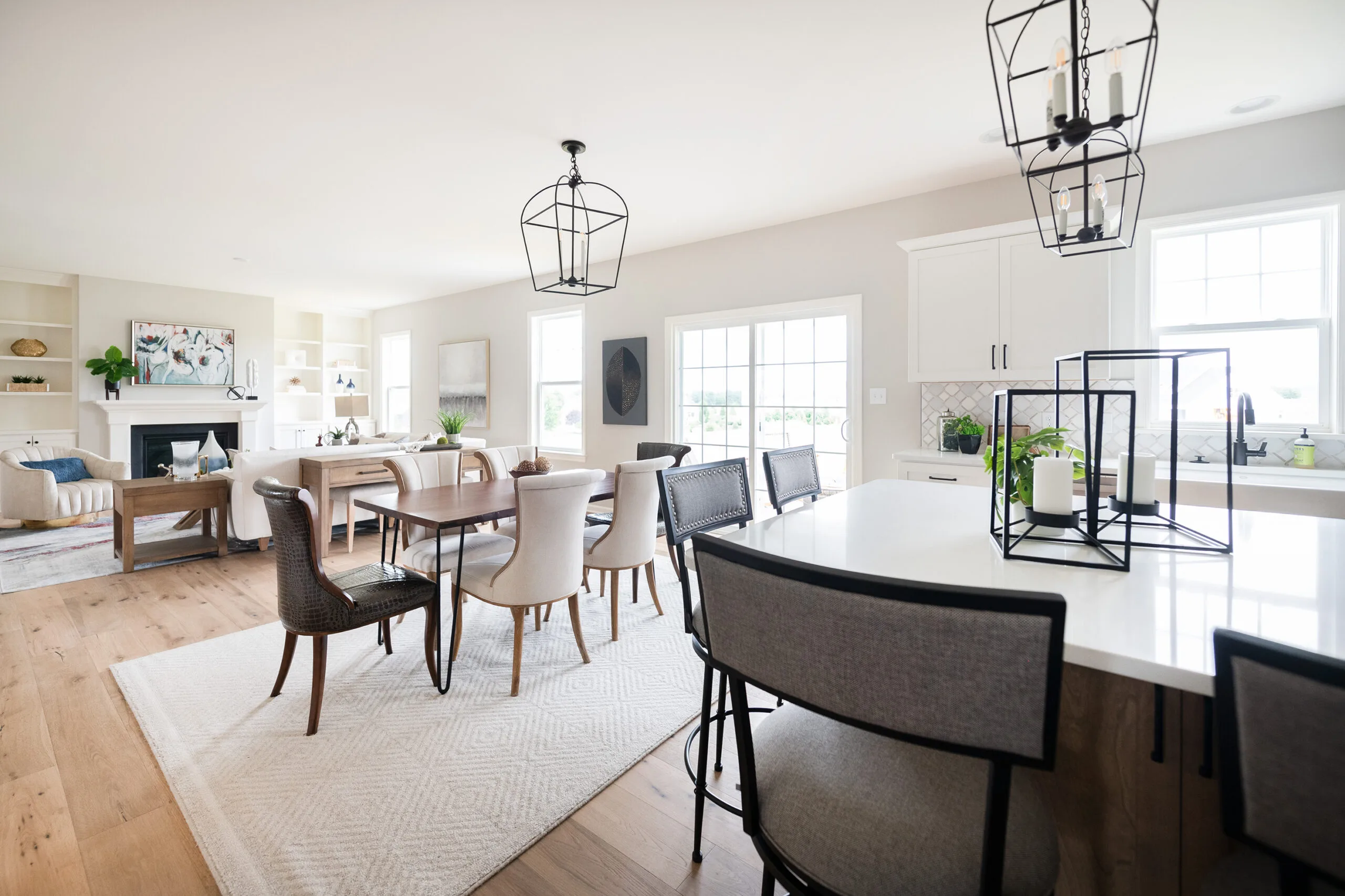 Bright, modern open-concept kitchen and dining area with neutral décor, white cabinetry, pendant lighting, a large island with stools, and a wooden dining table surrounded by upholstered chairs.