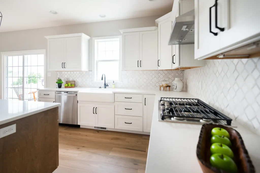 Modern kitchen with white cabinets, stainless steel appliances, a gas stove, and a large sink under a window. There are green apples in a wooden bowl on the counter and natural light fills the space.