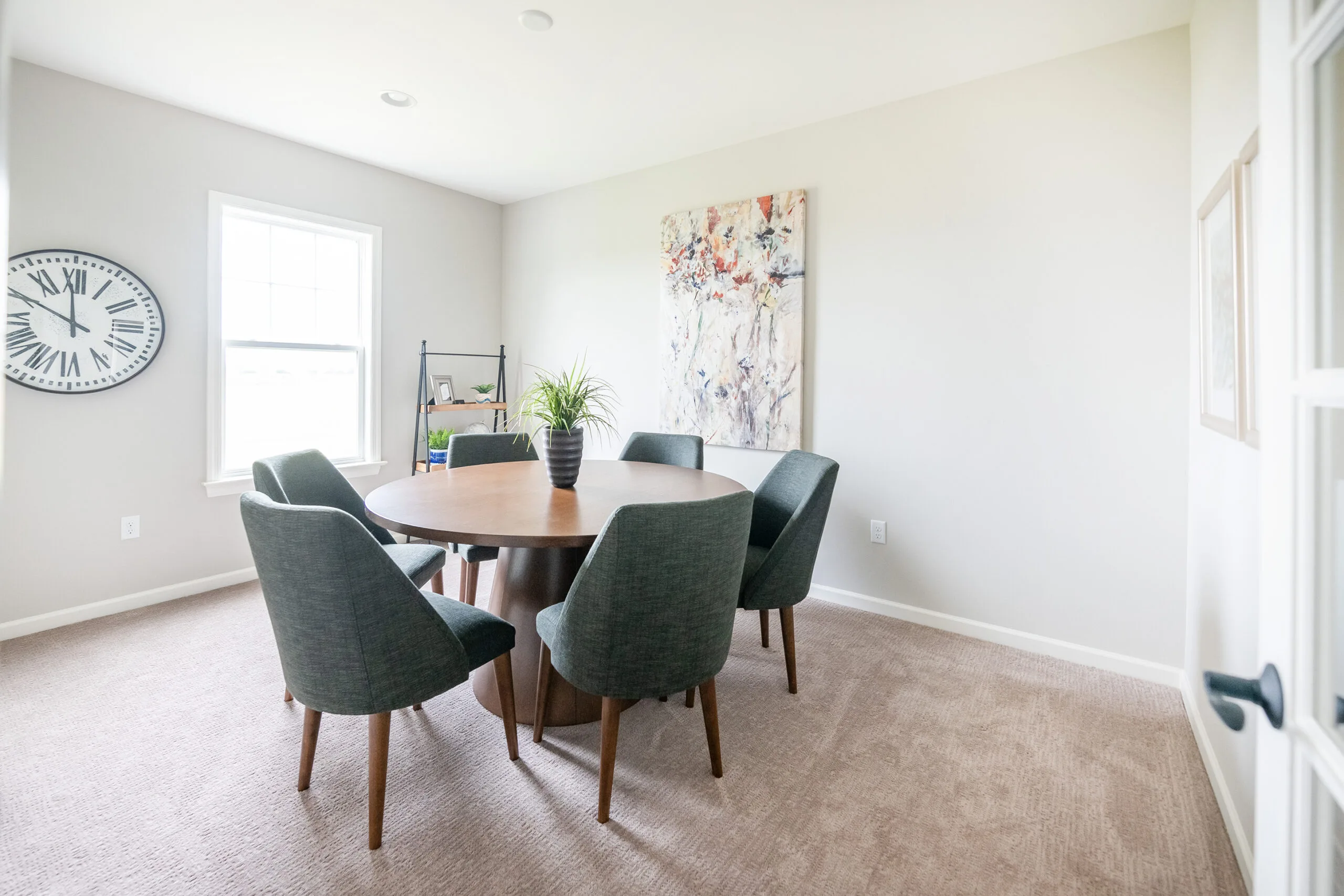 A bright, modern dining room with a round wooden table, six dark green chairs, a potted plant centerpiece, a large wall clock, abstract wall art, and beige carpet. Sunlight streams in through a window.