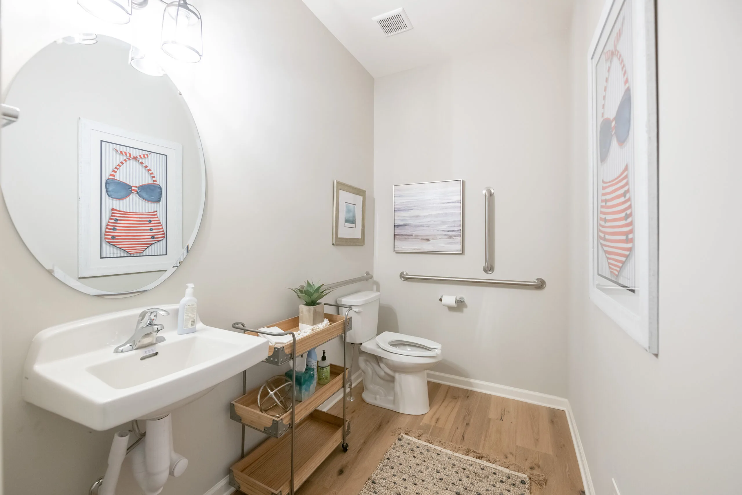 A bright, modern bathroom with a round mirror above a white sink, a toilet with safety grab bars, light wood flooring, and beach-themed artwork on the walls. A rolling cart holds toiletries and a framed photo.