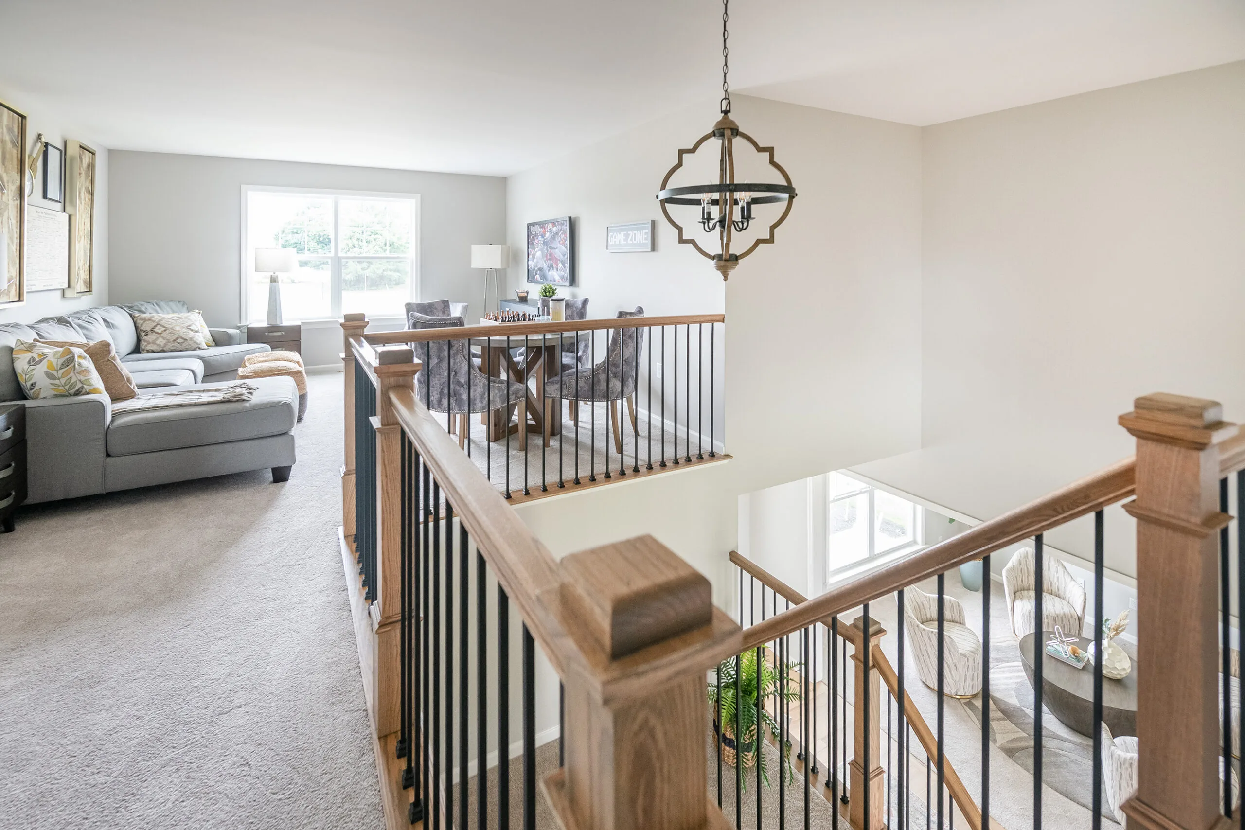 Modern, bright living space with beige carpeting, a gray sectional sofa, dining table with chairs, large windows, and a wood and metal stair railing; light fixture hangs above the staircase.