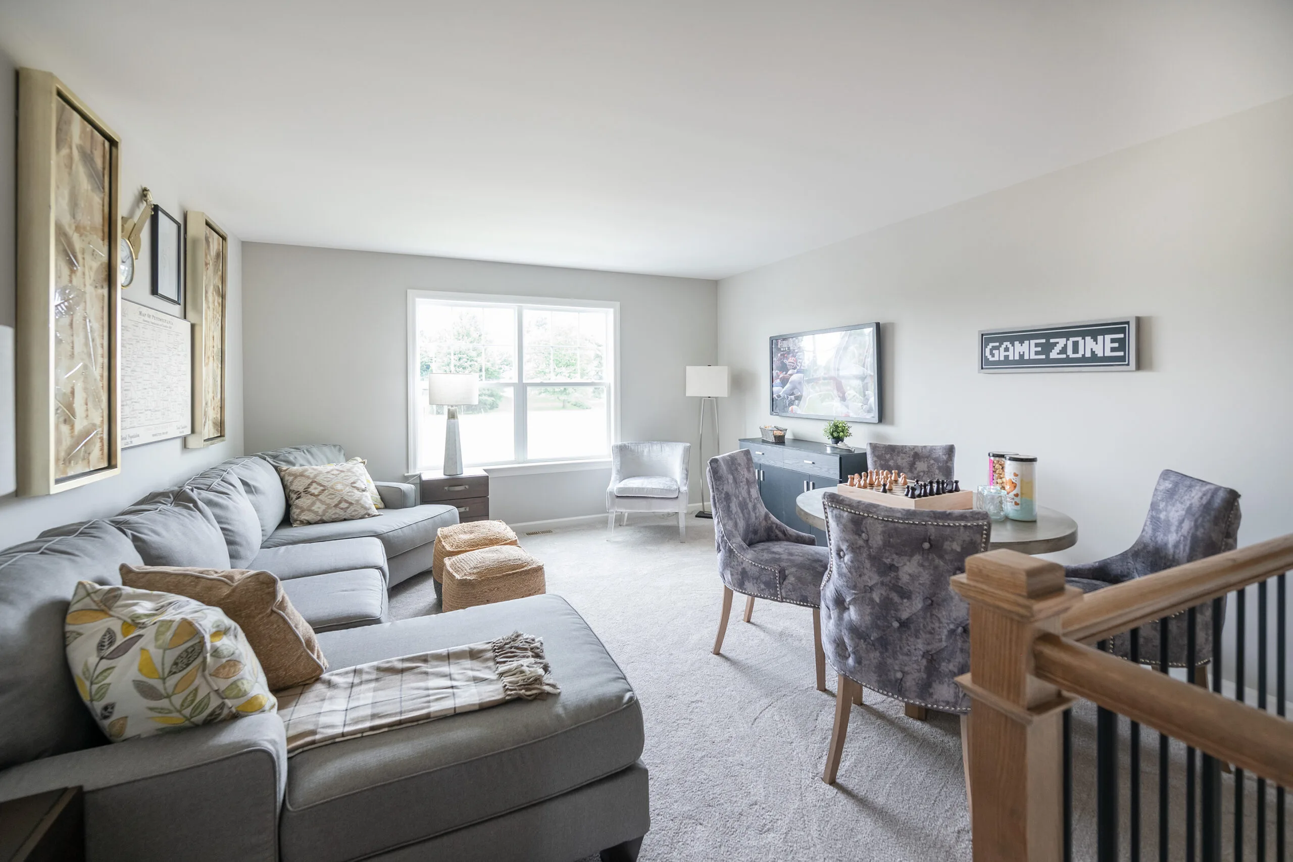 A bright living room with a large gray sectional sofa, two gray armchairs at a round table with a chessboard, a TV, “Game Zone” sign on the wall, and a window letting in natural light.