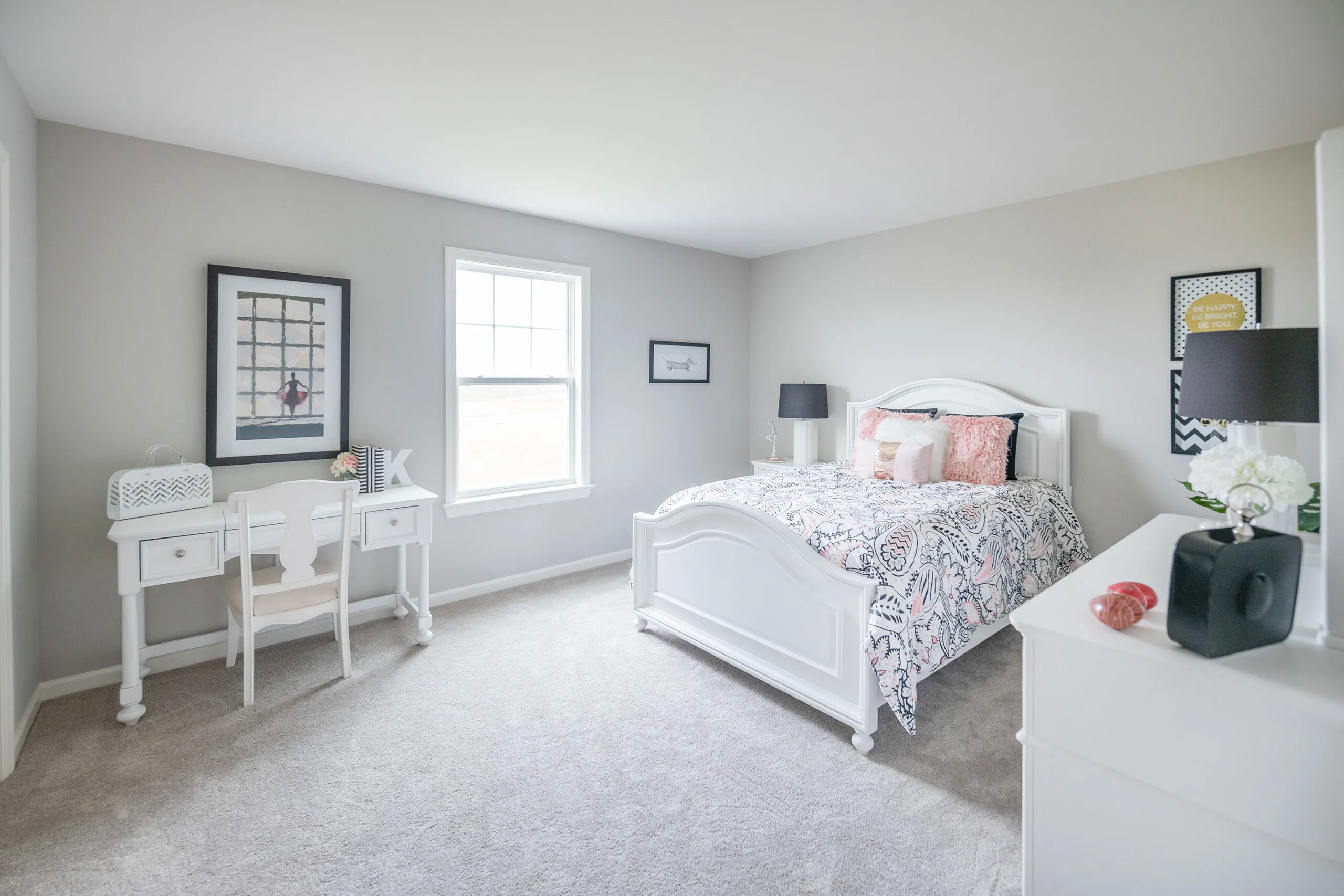 Bright bedroom with a white bed, patterned bedding, white desk and chair by a window, and simple wall art. Light gray walls and carpet create a clean, airy feel.